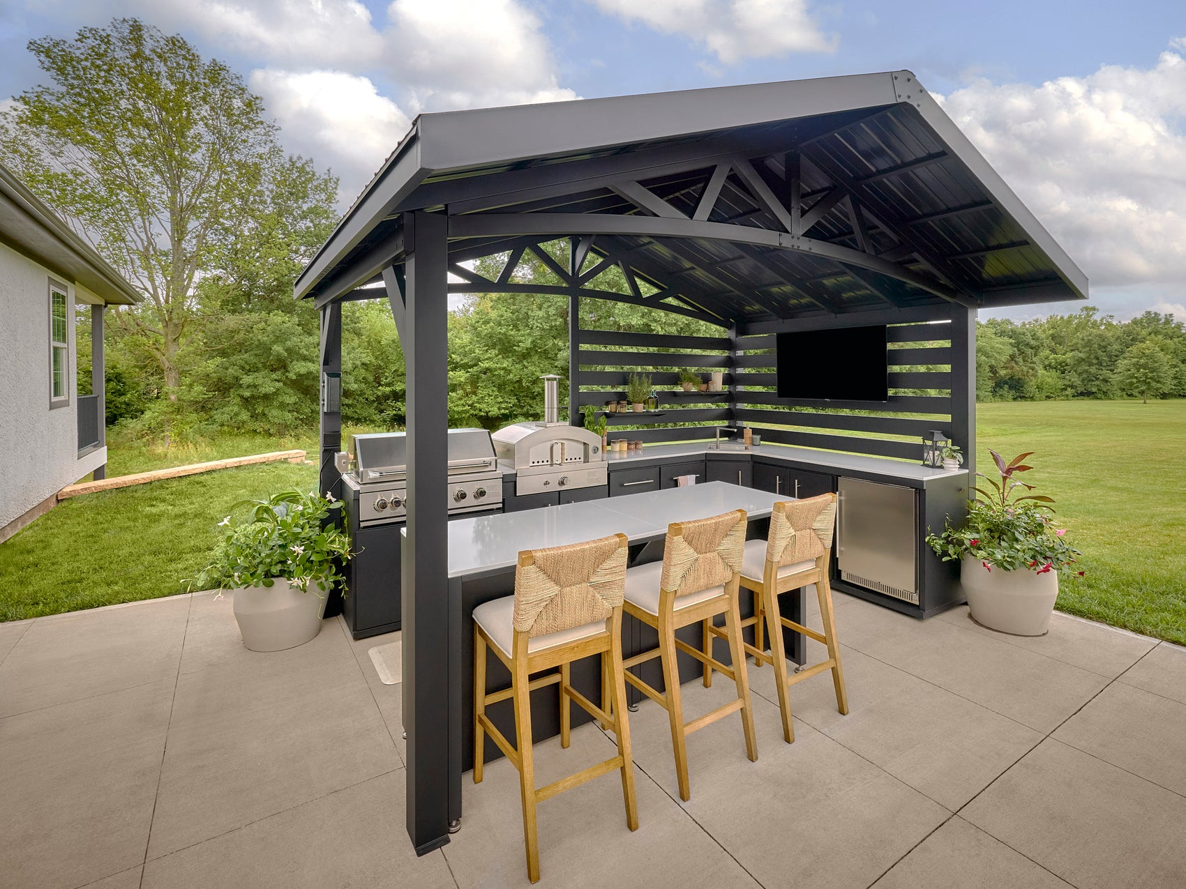 Steel pergola Langston patio kitchen with built-in grill, bar seating, and countertops overlooking green lawn and trees.