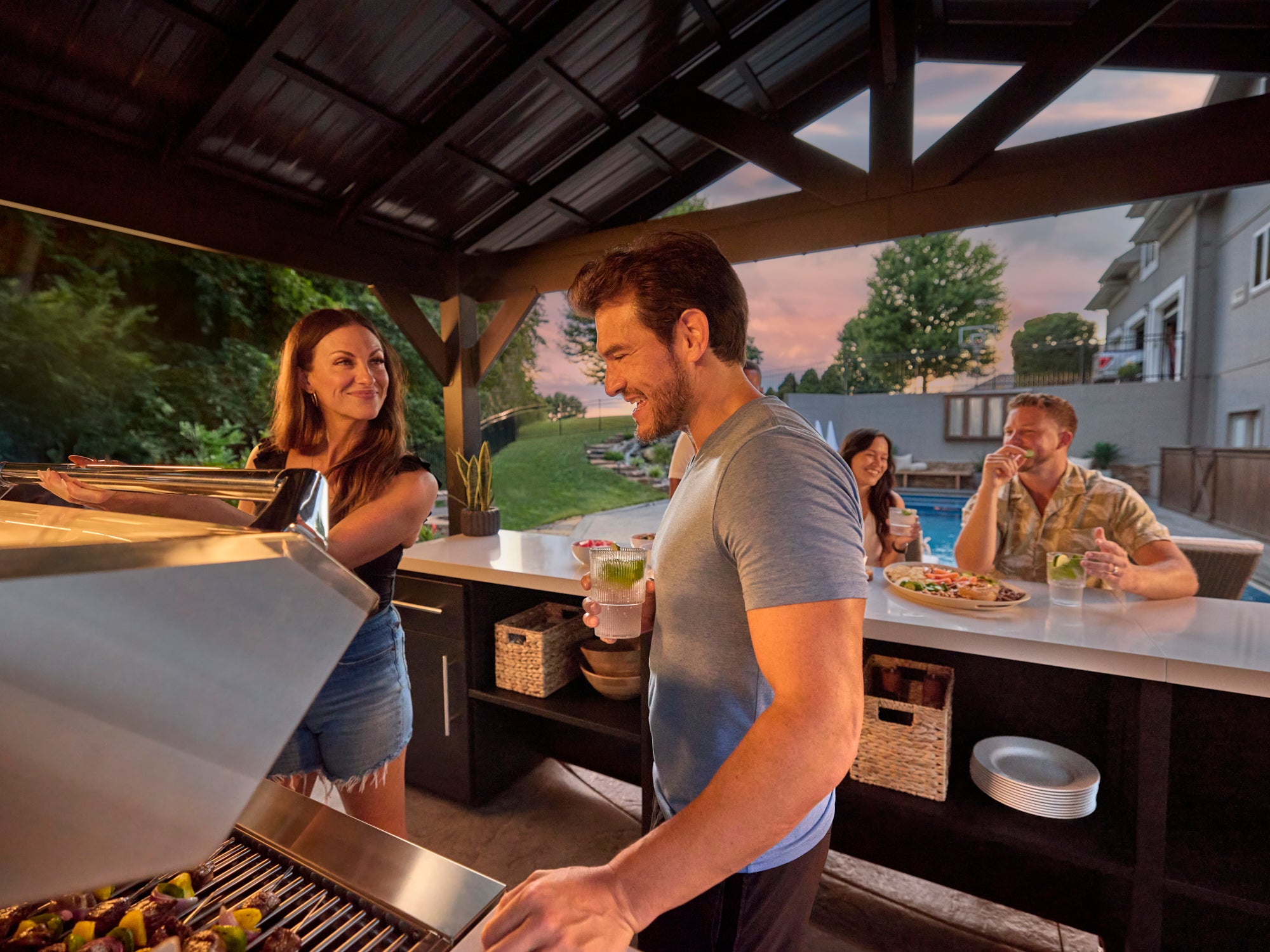 Range Outdoor Living Lancaster kitchen bar, guests entertaining by pool at sunset in modern backyard.