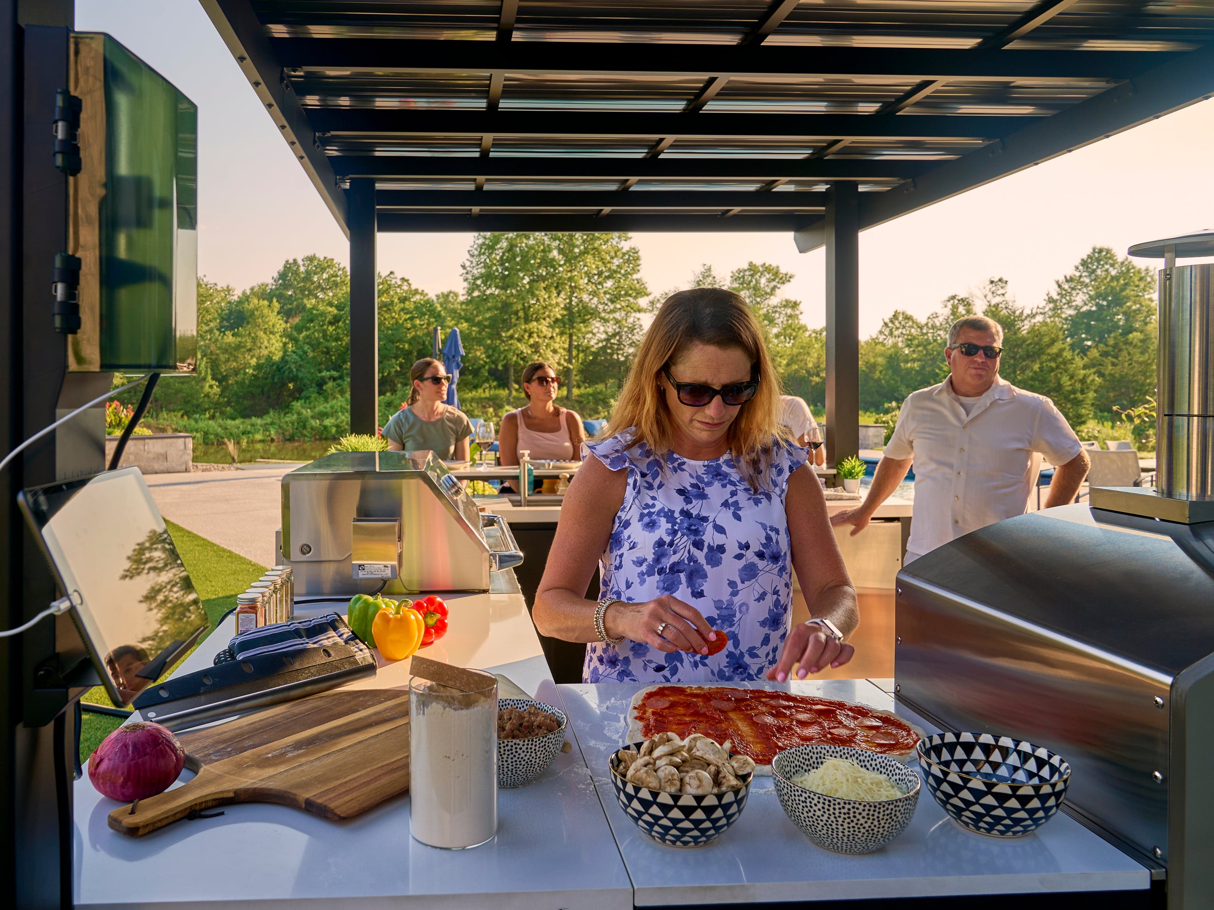 Family gathering at Edgemont Bay outdoor kitchen island with woman prepping pizza on engineered stone counter.
