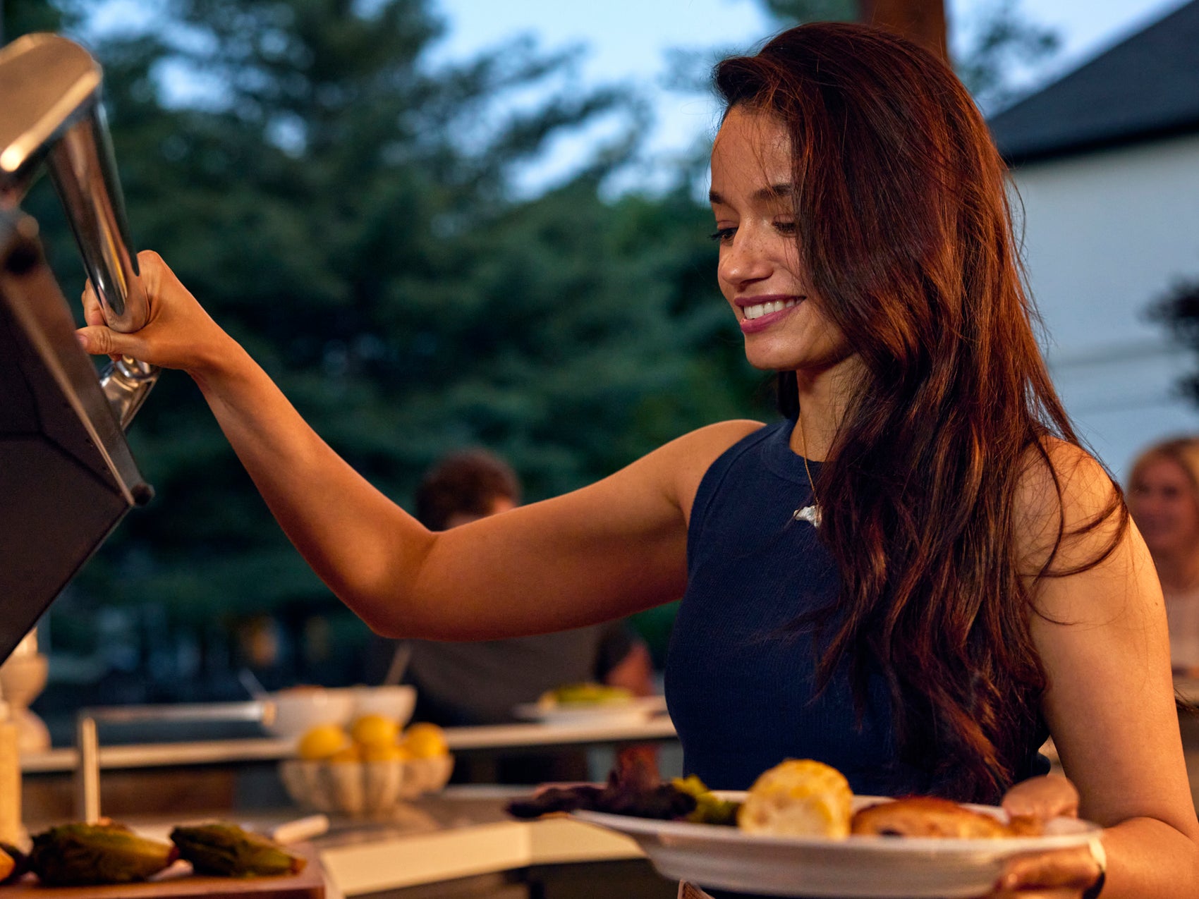 Woman grilling at outdoor dinner party under steel roof of Emory kitchen, showcasing backyard pergola ideas.
