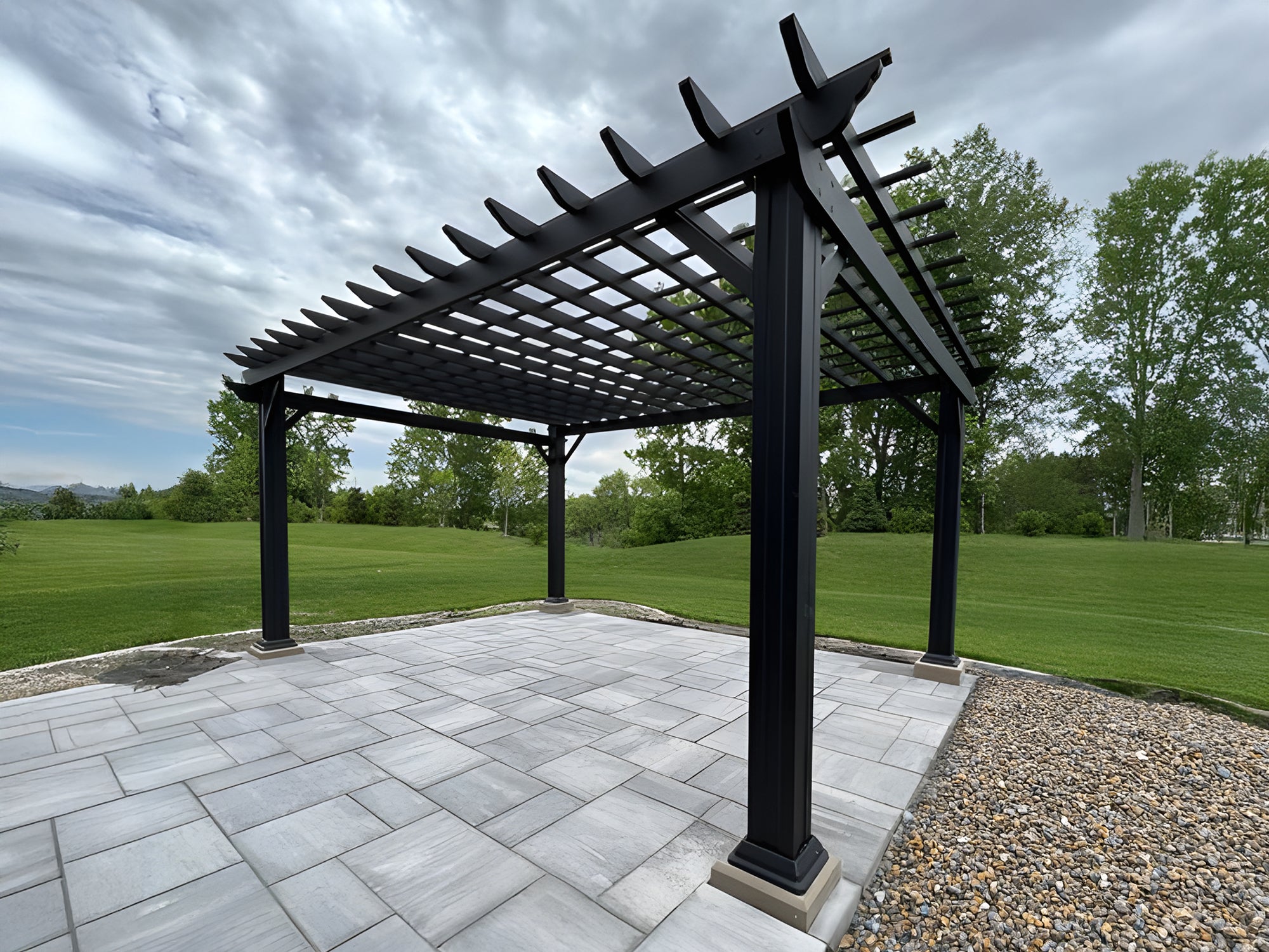 Black Stratford steel pergola with trellis roof over gray paver patio in open lawn with trees and dramatic cloudy sky.