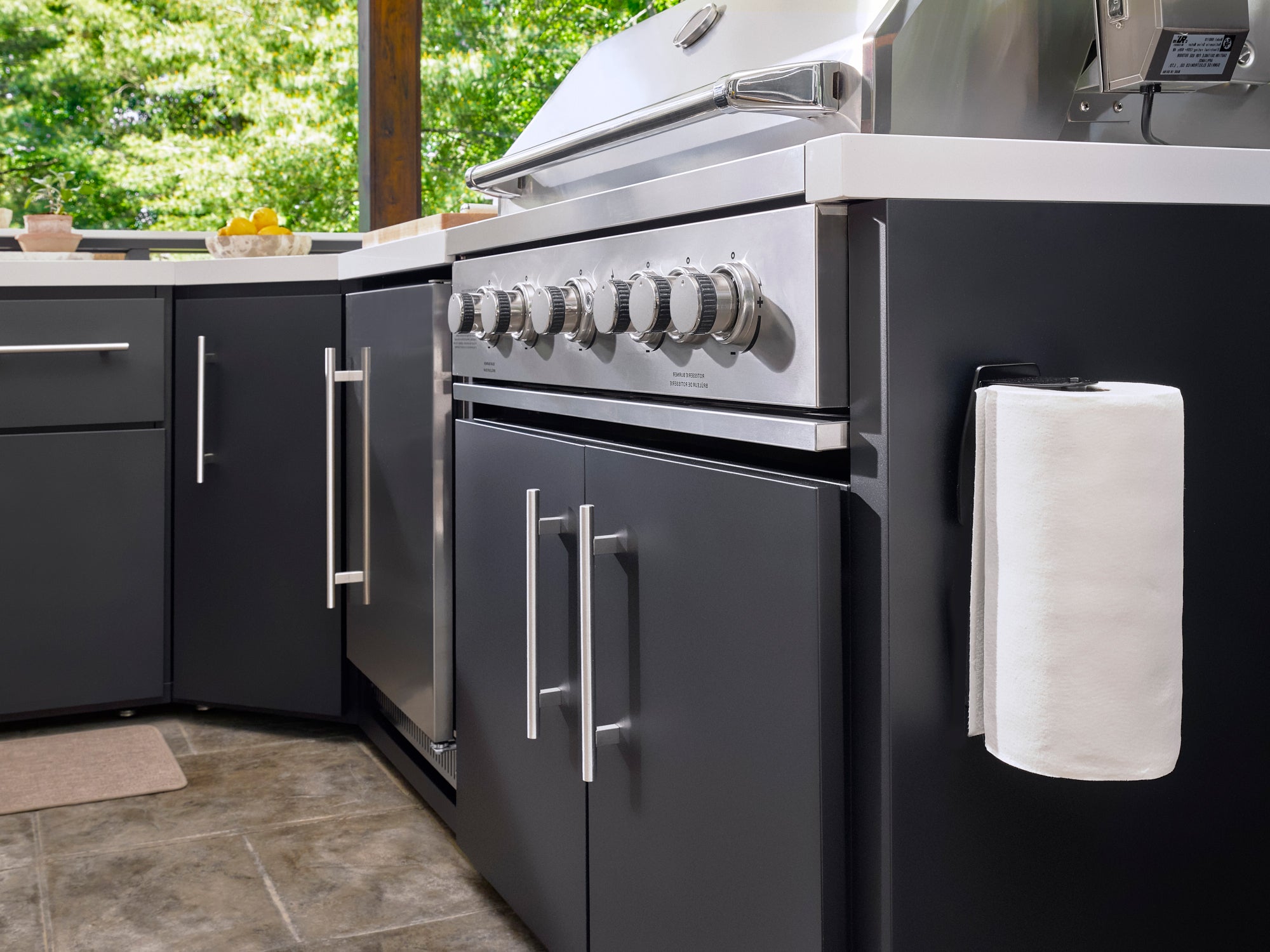 Emory black outdoor kitchen with built-in grill and paper towel holder beneath patio pergola with lush greenery background.