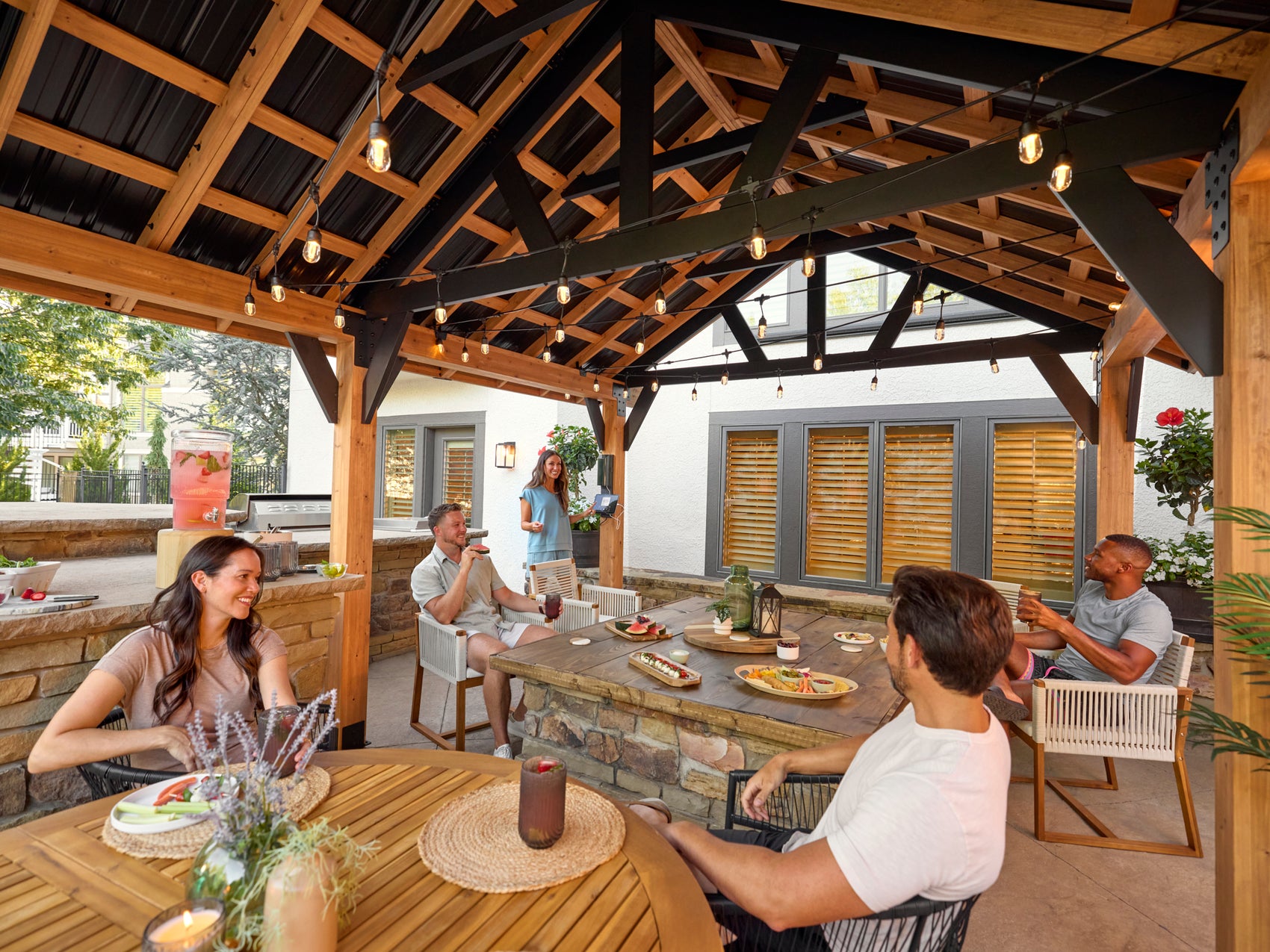 People dining under Lodge outdoor gazebo with exposed timber beams, string lights, and stone kitchen island on patio.