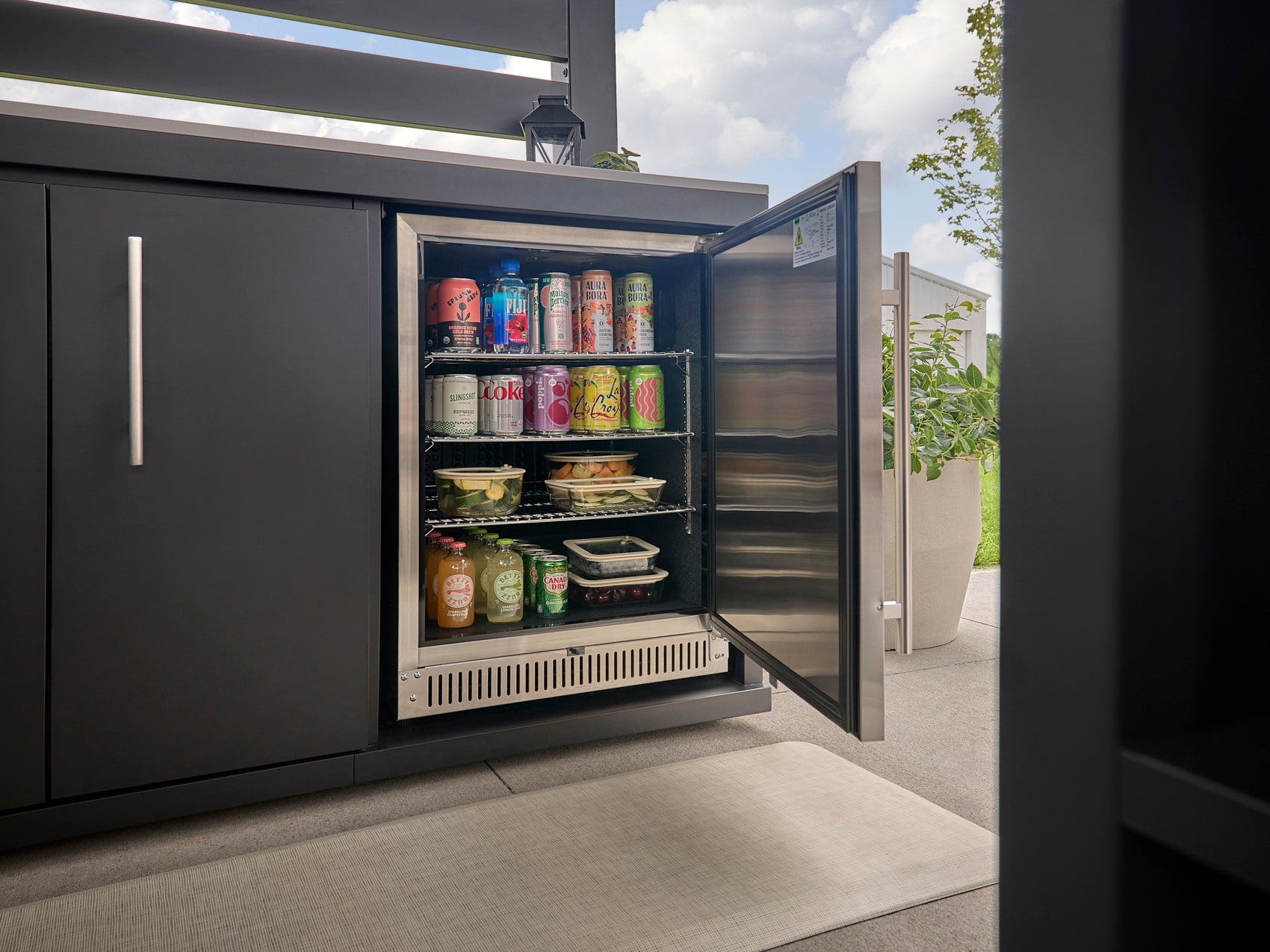 Built-in outdoor fridge with open door displaying beverages and food containers on wire shelves in modern kitchen setting.