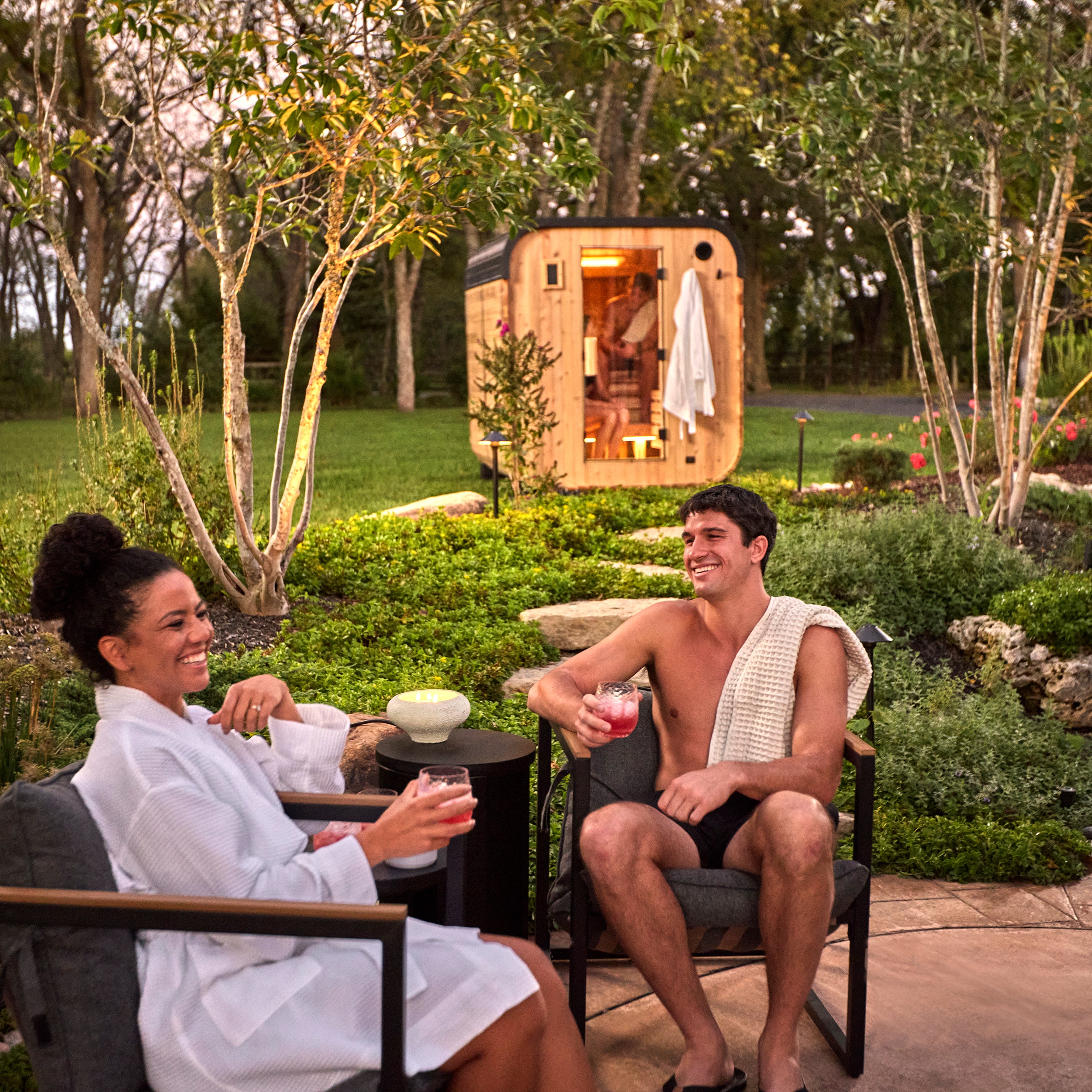 Couple enjoying drinks on patio with illuminated Lennon cube cedar backyard sauna at dusk