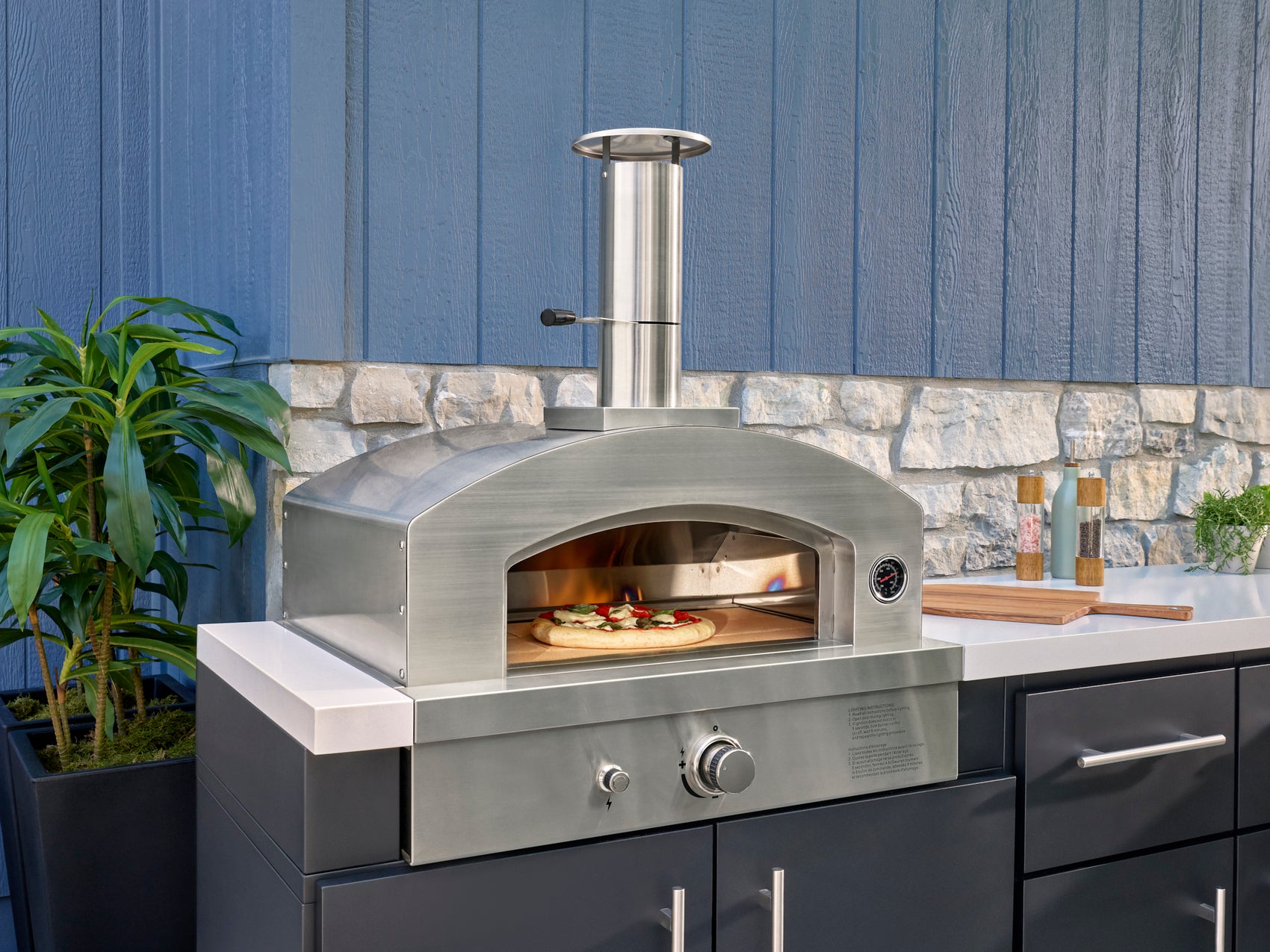 Stainless steel outdoor pizza oven with arched door, thermometer, control knobs, and chimney on engineered stone countertop.