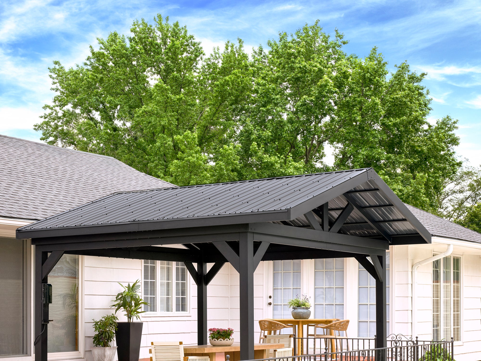 Black steel Lancaster gazebo with roof over patio dining area with decorative braces against white home and green trees.