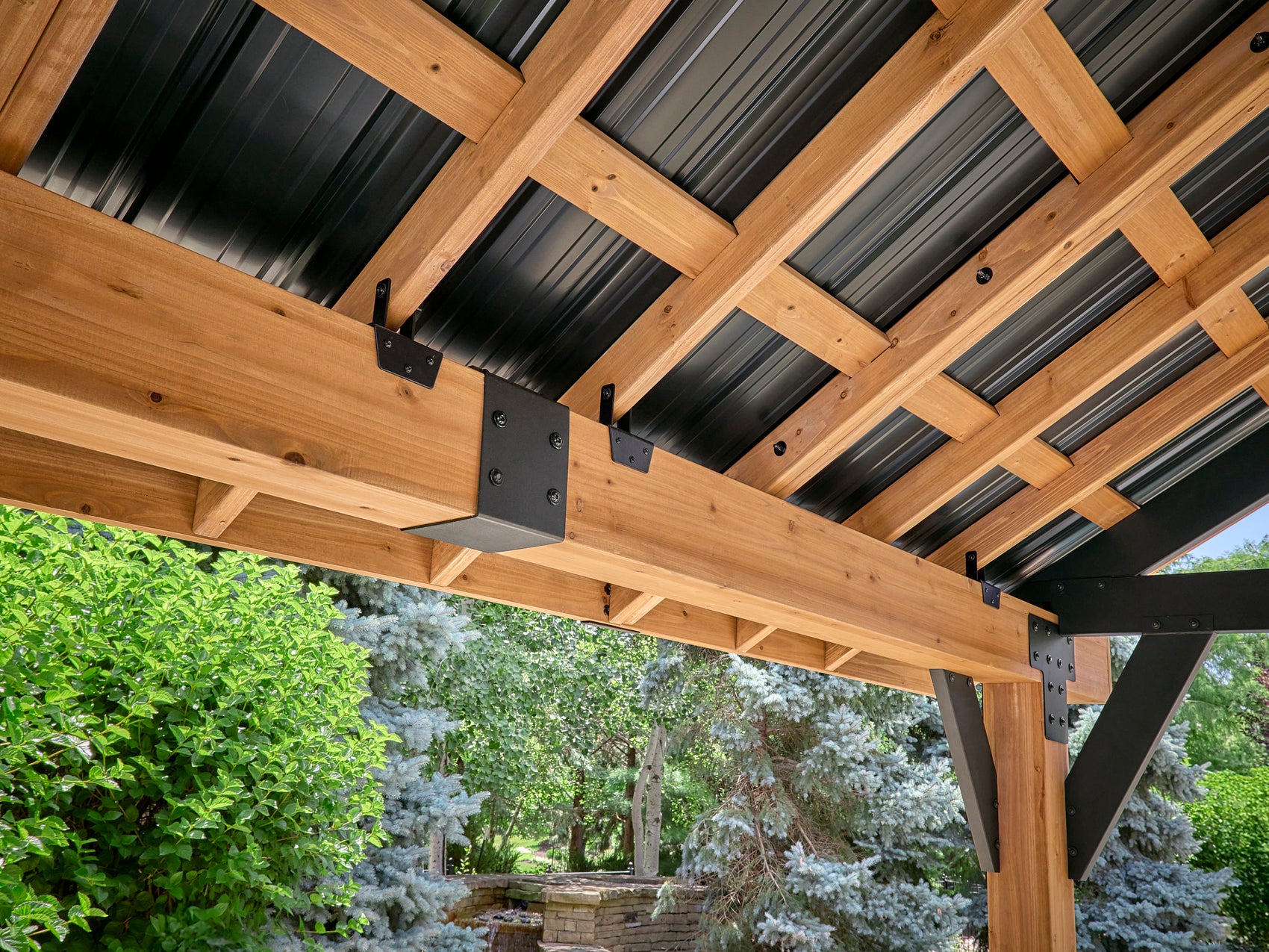 Metal bracket connecting timber beam to steel post on Lodge hardtop gazebo with corrugated roof panels and garden view.