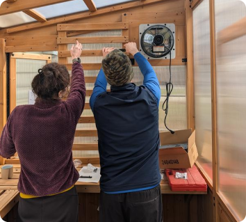 Two people working on a wooden structure with a fan attached, in a workshop setting.