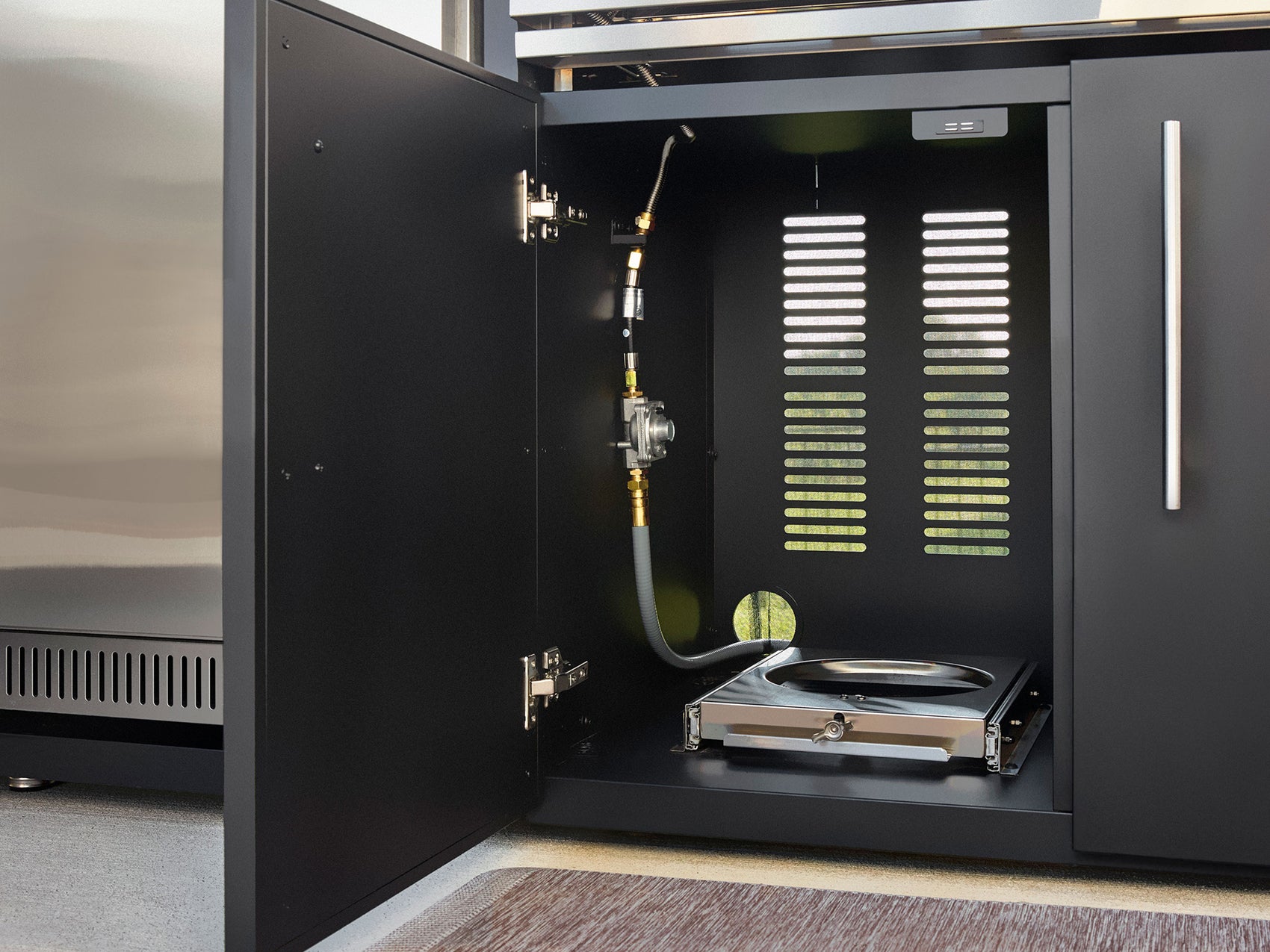 Open outdoor kitchen cabinet revealing gas line hookup with regulator, flexible hose, and drip tray with ventilation slots.