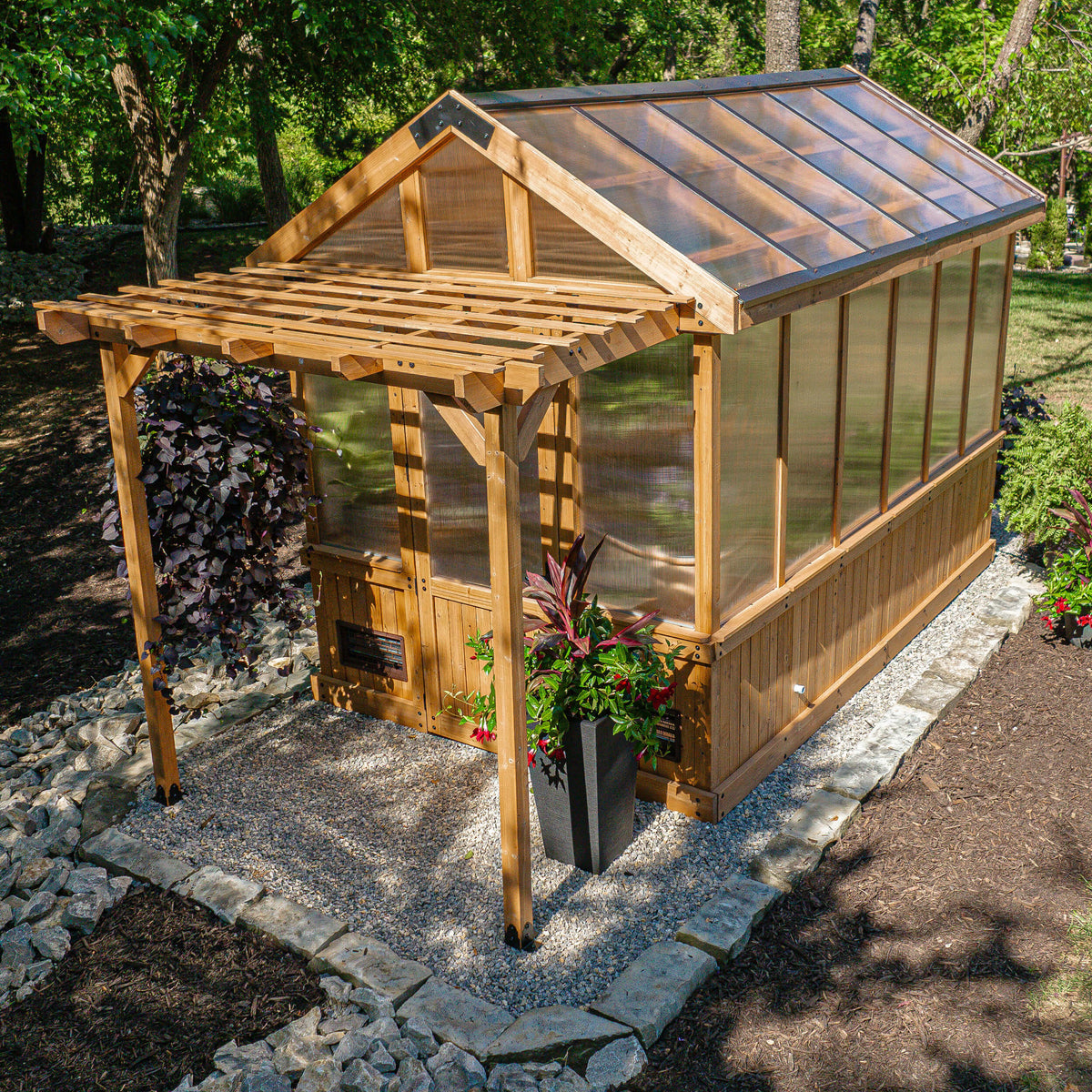 Bellerose wooden greenhouse with a polycarbonate roof and walls panels, surrounded by plants and trees.