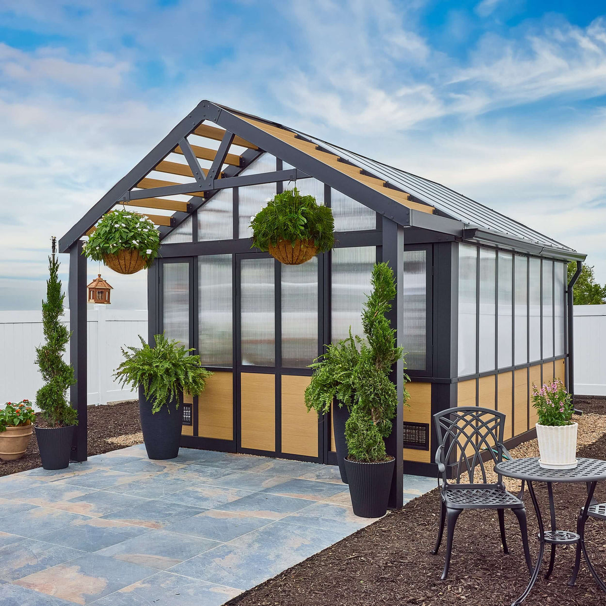 A modern greenhouse with a dark gray frame and light wood paneling, set outdoors on a stone patio with potted plants, a small patio table, and a white fence under a blue sky.