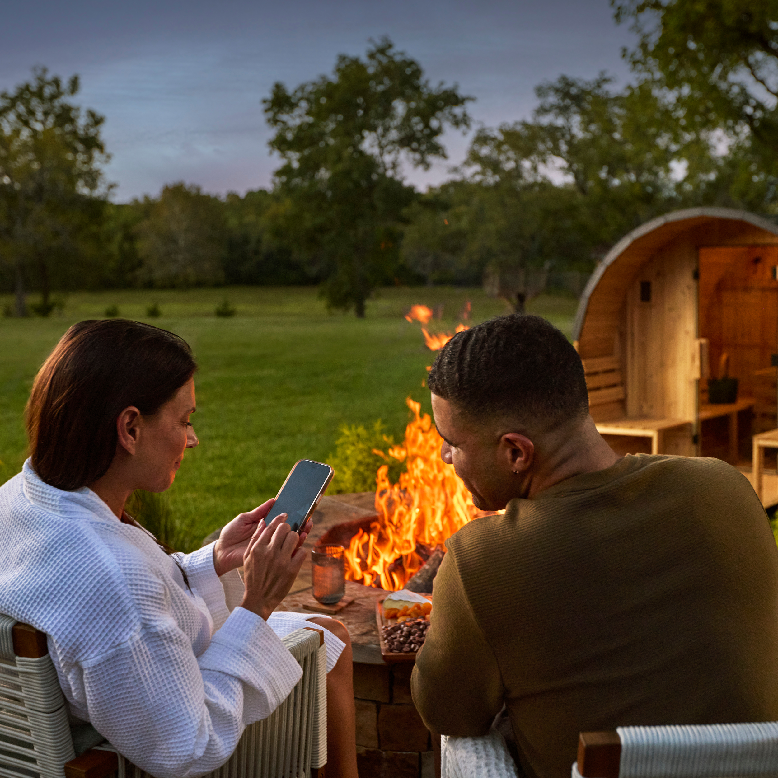 Two people sitting by a fire pit outdoors, using a smartphone.