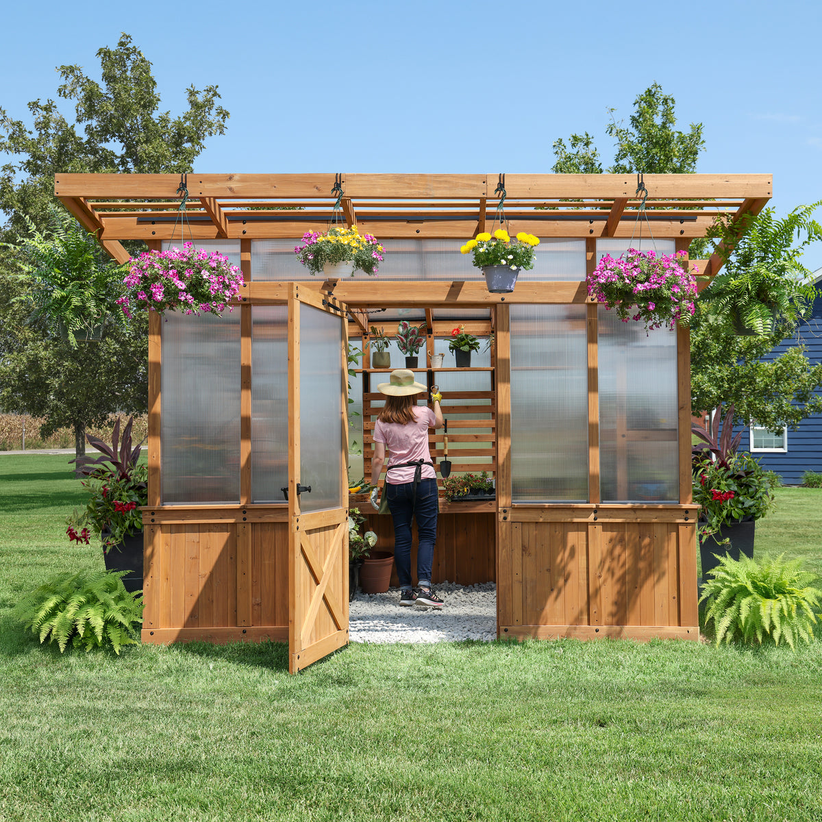 Woman tending plants inside the 11x7 Poppy cedar and polycarbonate greenhouse with an integrated pergola overhang.