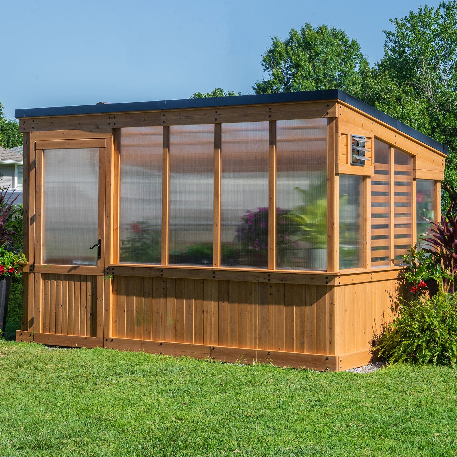 A cedar and polycarbonate Zalie greenhouse with a wide entry door, large front panels, and a side intake vent, set on a lawn.