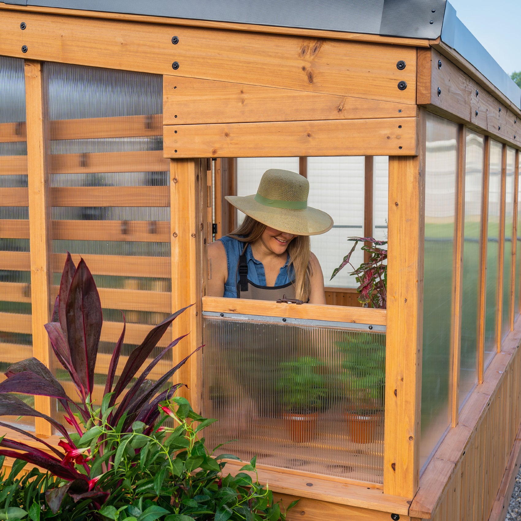 Woman tending plants inside the Zalie outdoor greenhouse, viewed through an open side window.