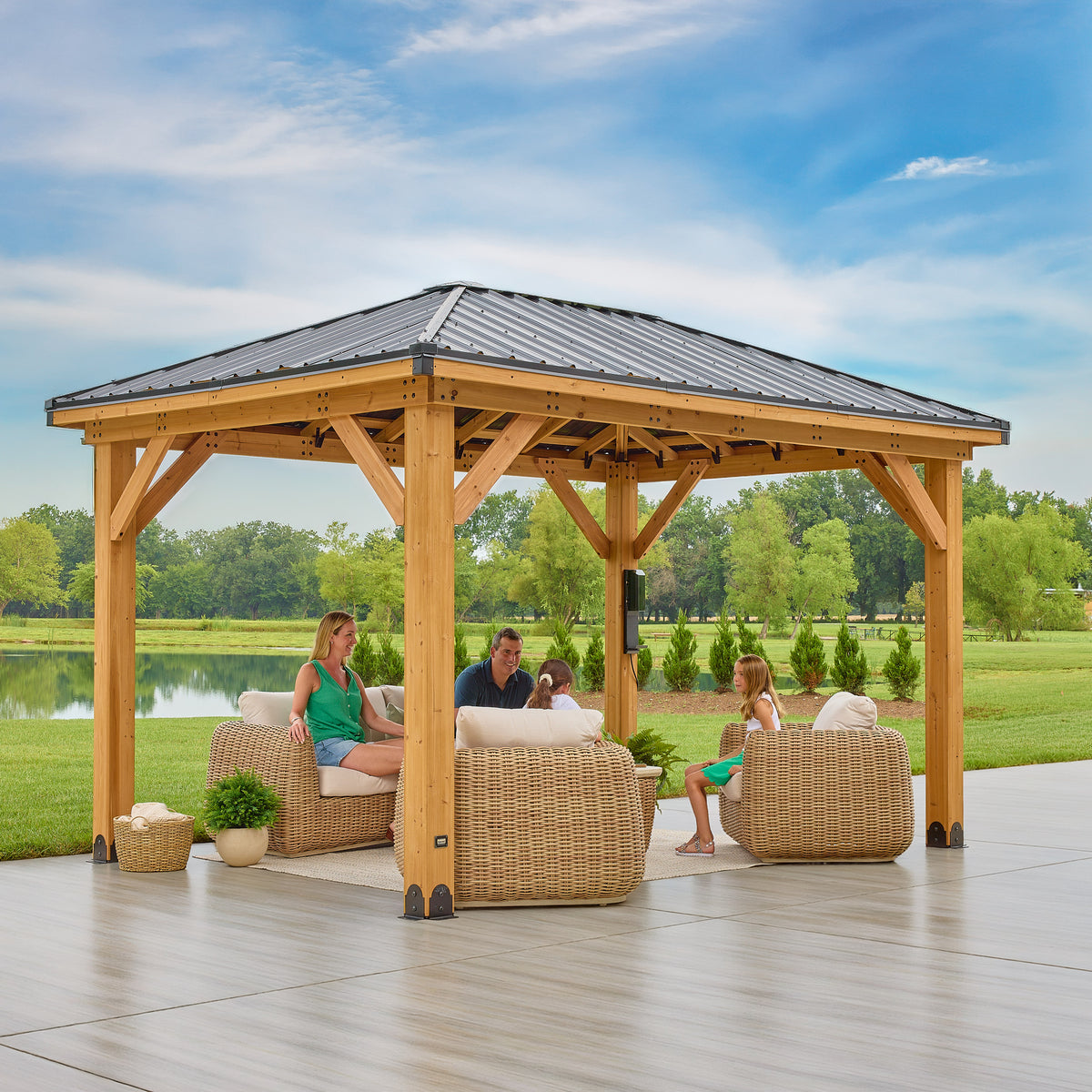 Cedar posts and black steel hip roof sheltering a family seated beneath a hardtop gazebo on a paved patio.