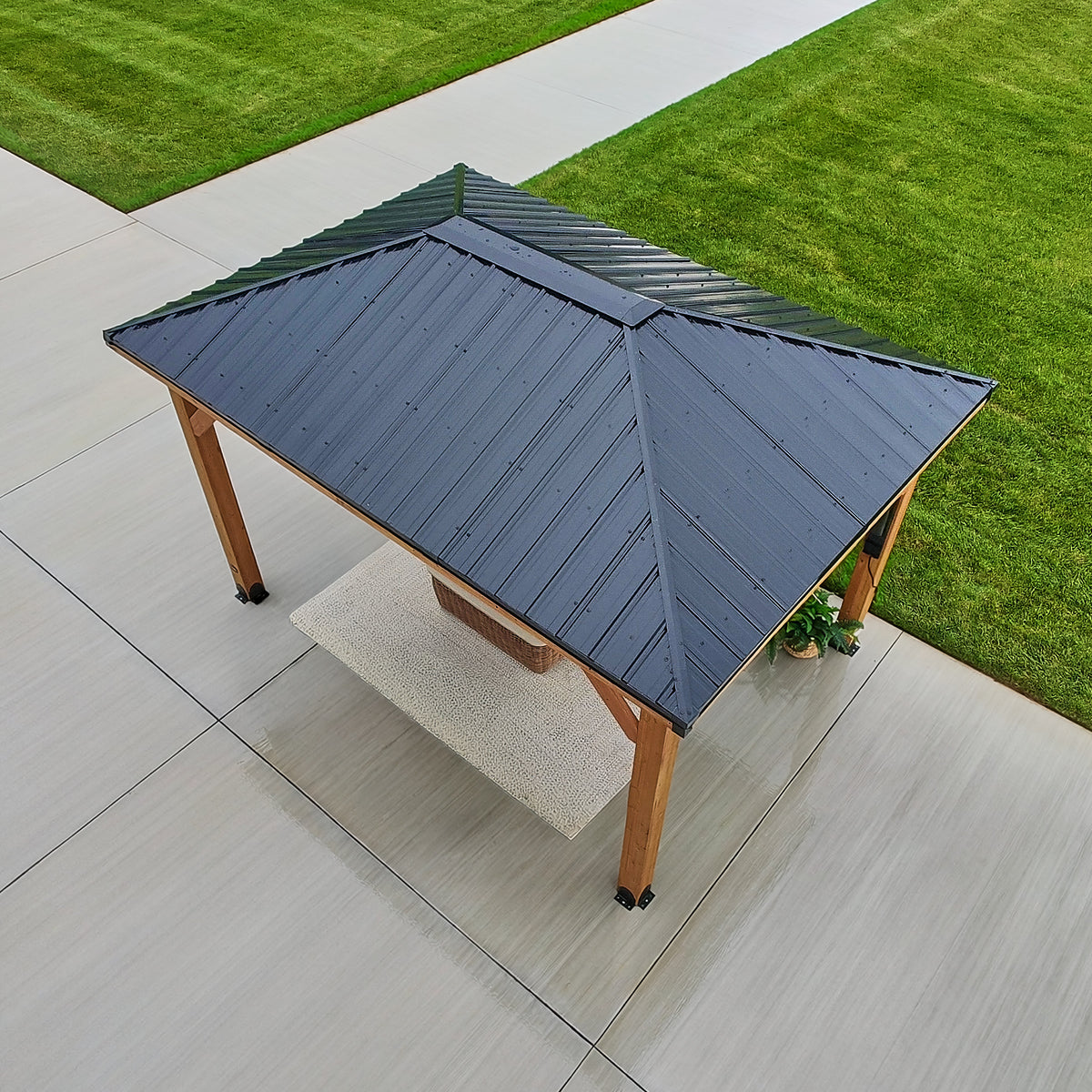 Black steel hip roof of a metal roof gazebo angled to four sides above cedar posts on a paved patio.