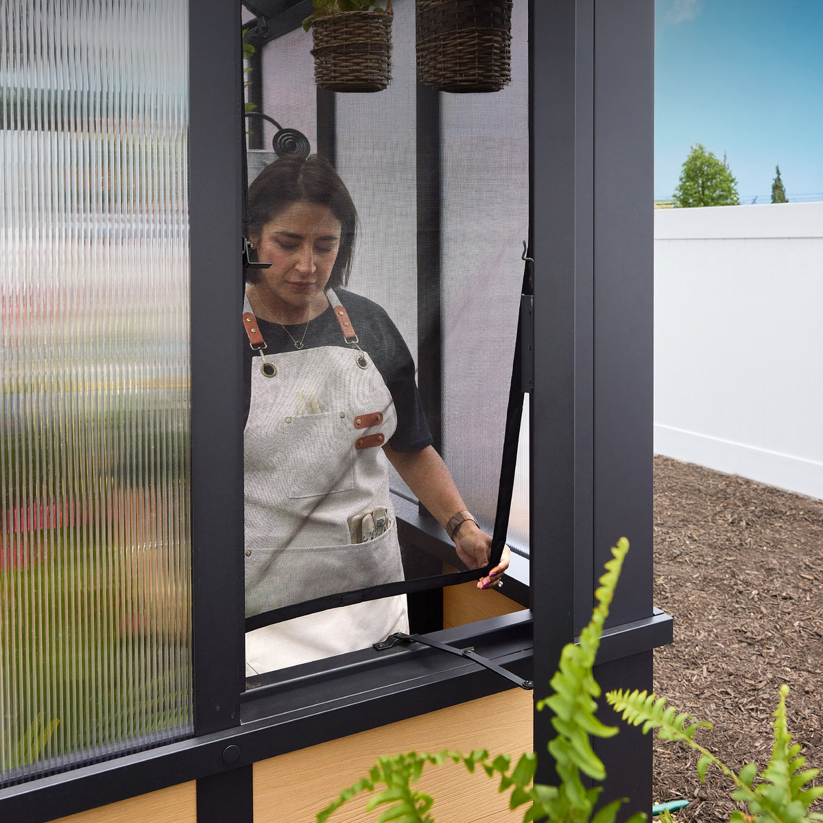 Woman opening an operable window on a walk-in powder-coated steel greenhouse for passive ventilation.