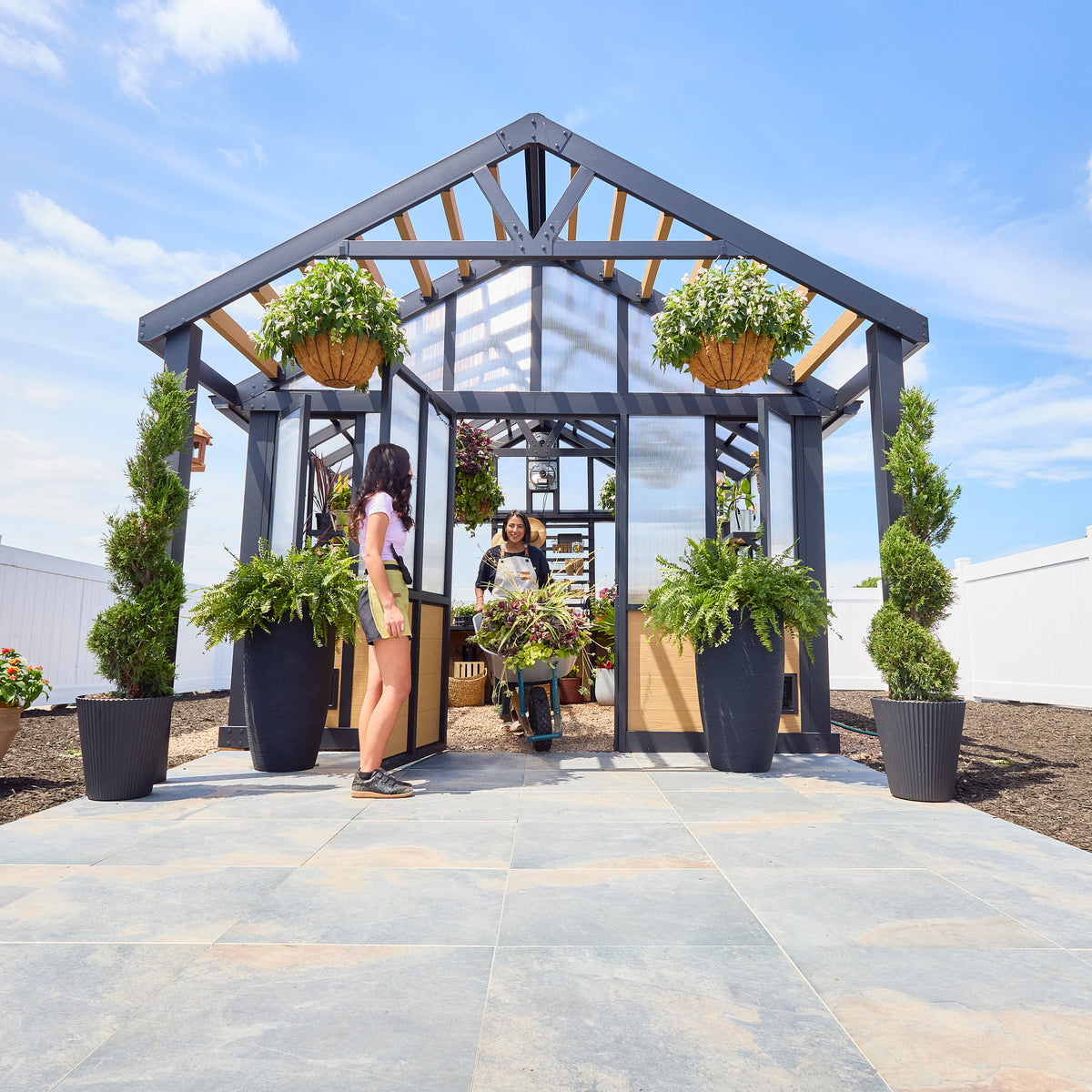 Two women at the open wide entry door of a DIY powder-coated steel greenhouse with potted plants on the shelves inside.