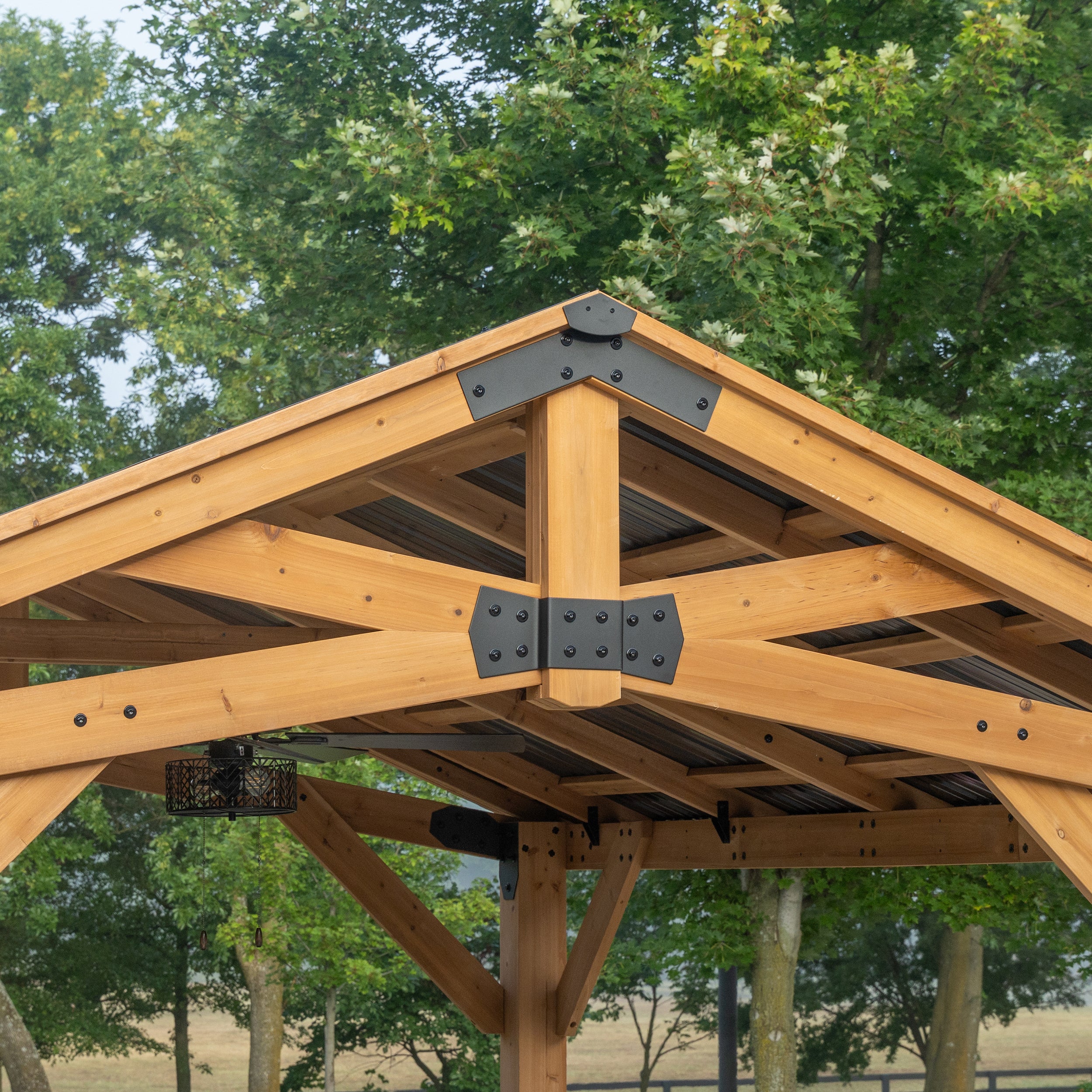 Peak ridge bracket of galvanized steel gazebo connecting cedar rafters and large post against tree canopy.