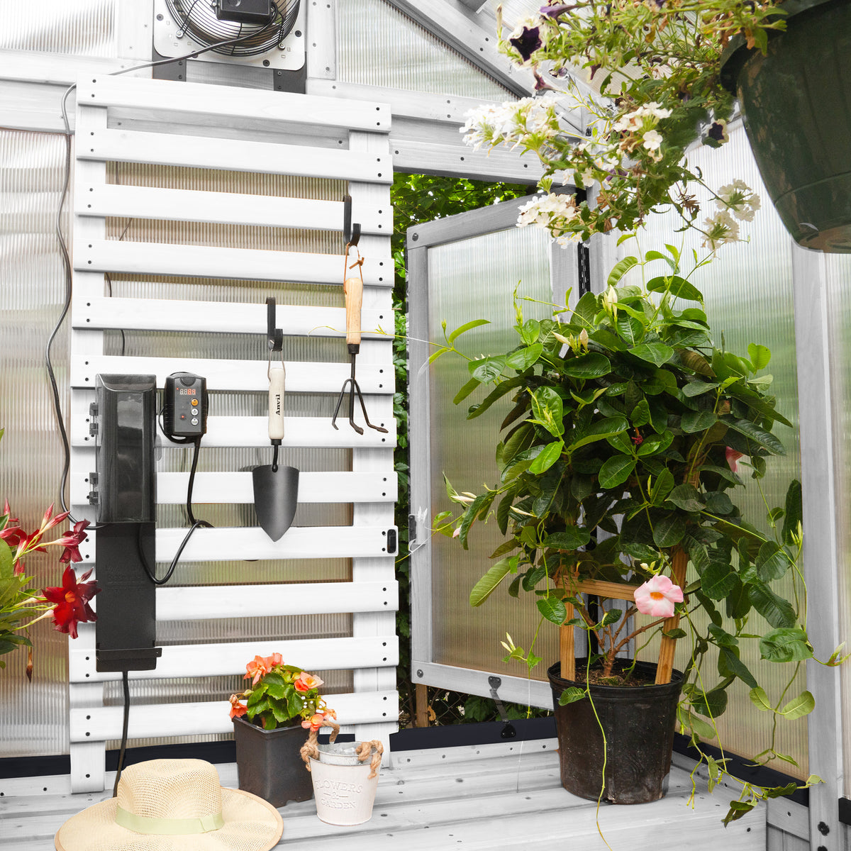 Slat wall with hanging garden tools, a heater with thermostat, and potted flowering plants inside a white-frame greenhouse.