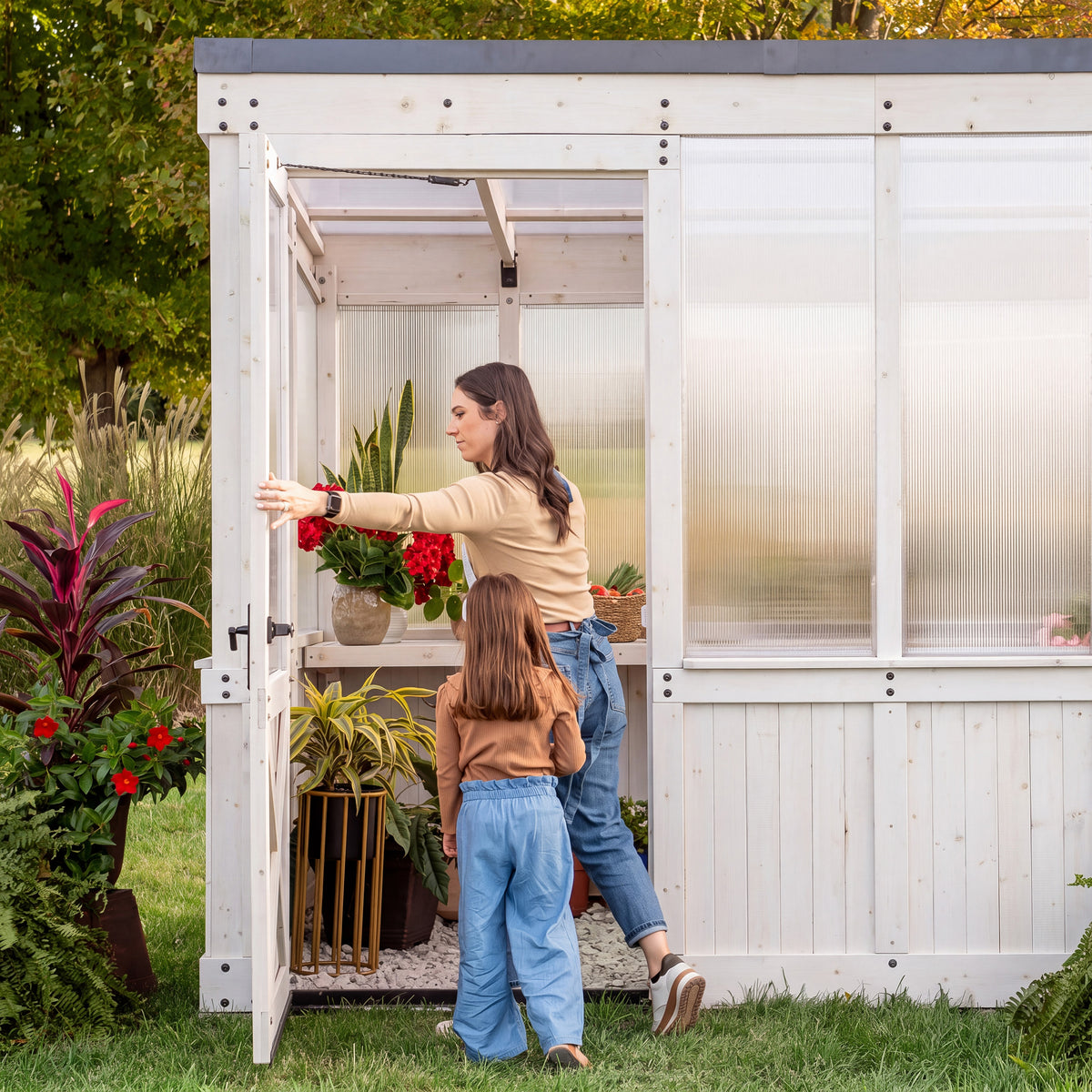 Woman and child at the open wide entry door of a garden white-frame greenhouse with potted plants on the shelves inside.