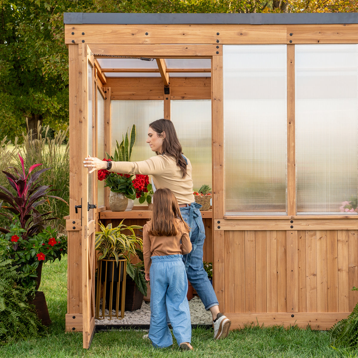 Woman and child at the open wide entry door of a backyard cedar greenhouse with potted plants on the shelves inside.