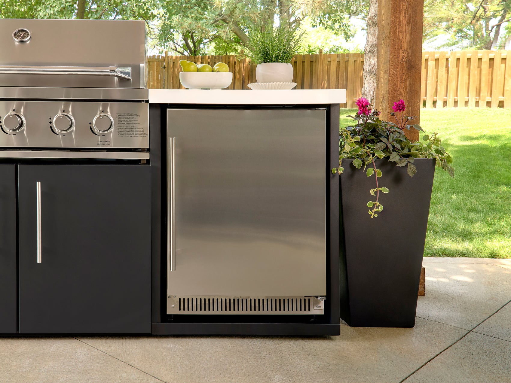 Outdoor refrigerator with stainless steel door built into cabinetry with white countertop, adjacent to grill and planter.