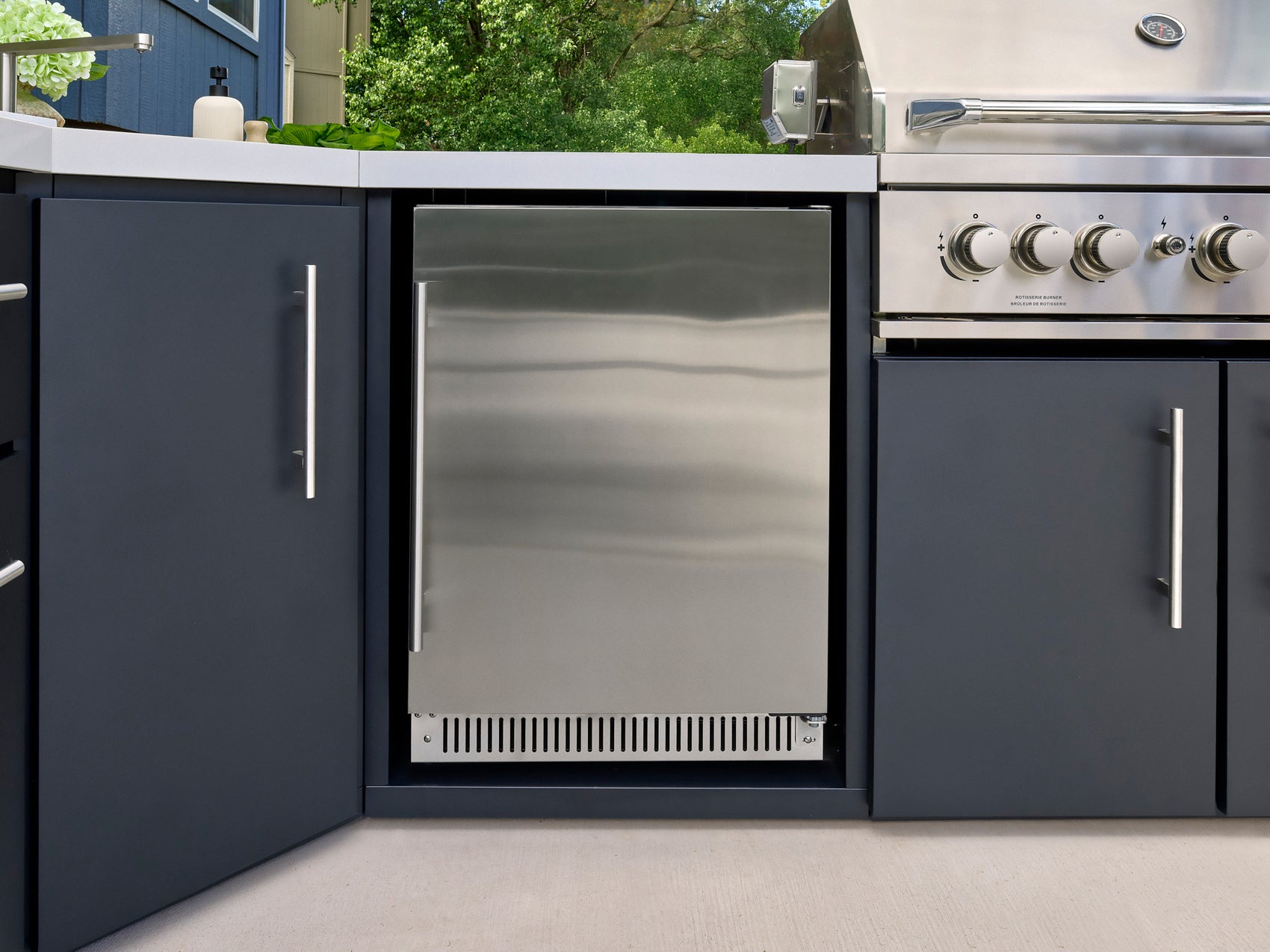 Stainless steel outdoor mini fridge built into dark gray cabinetry with white countertop and grill control knobs visible.