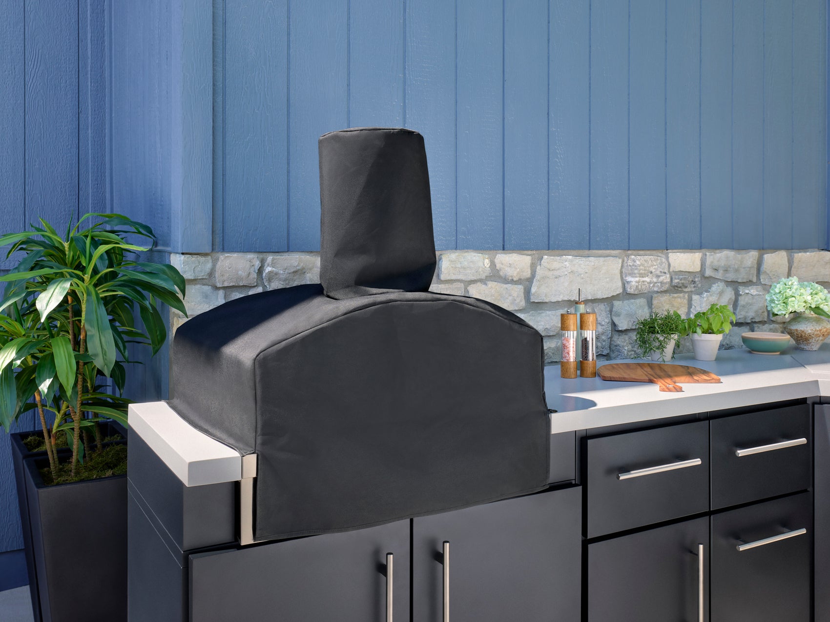 Covered built-in pizza oven on Emory outdoor kitchen counter with stone backsplash, plants, and cooking accessories nearby.