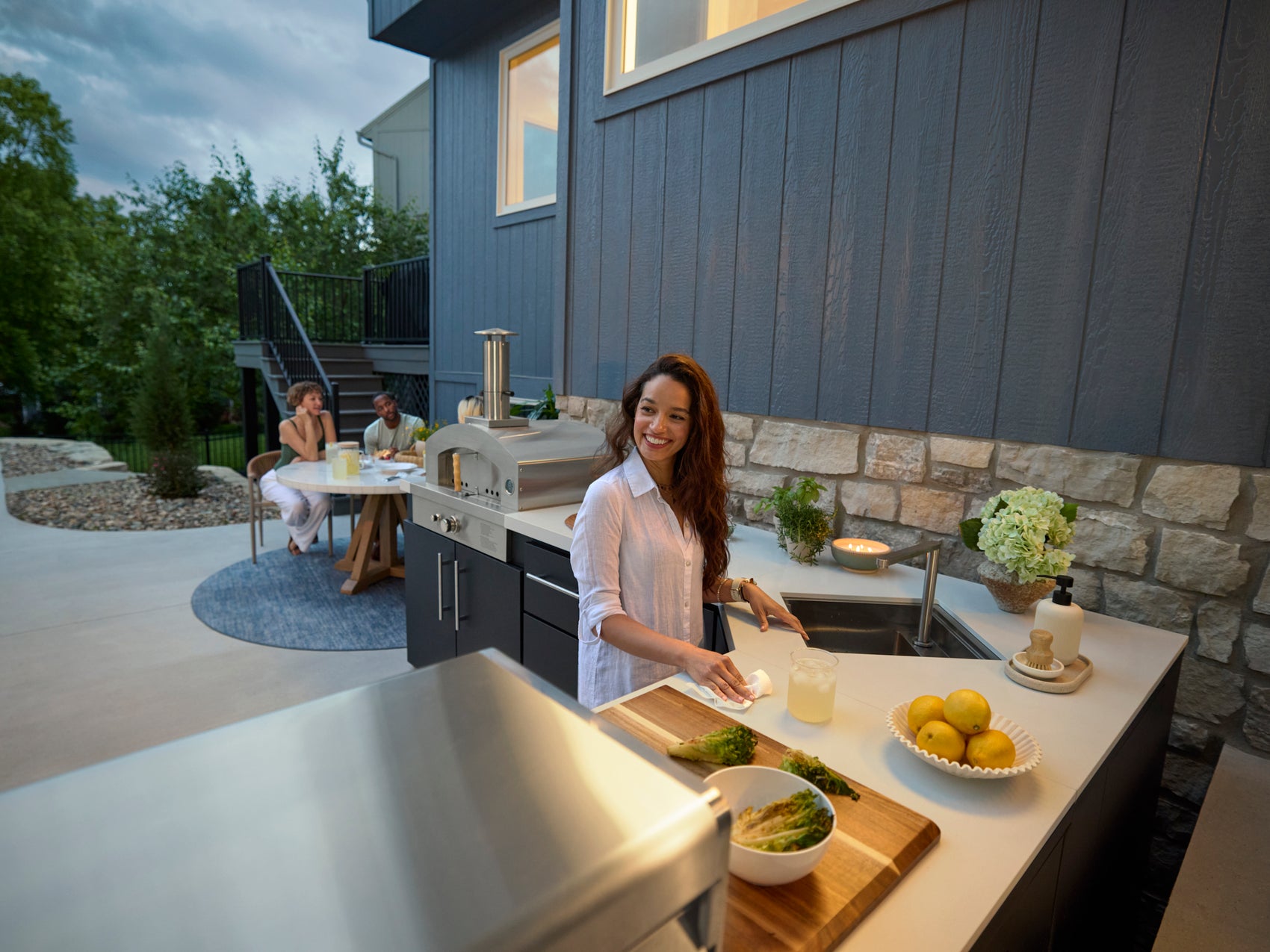 Woman preparing drinks at Emory outdoor kitchen island with pizza oven while guests relax at table near modern home.