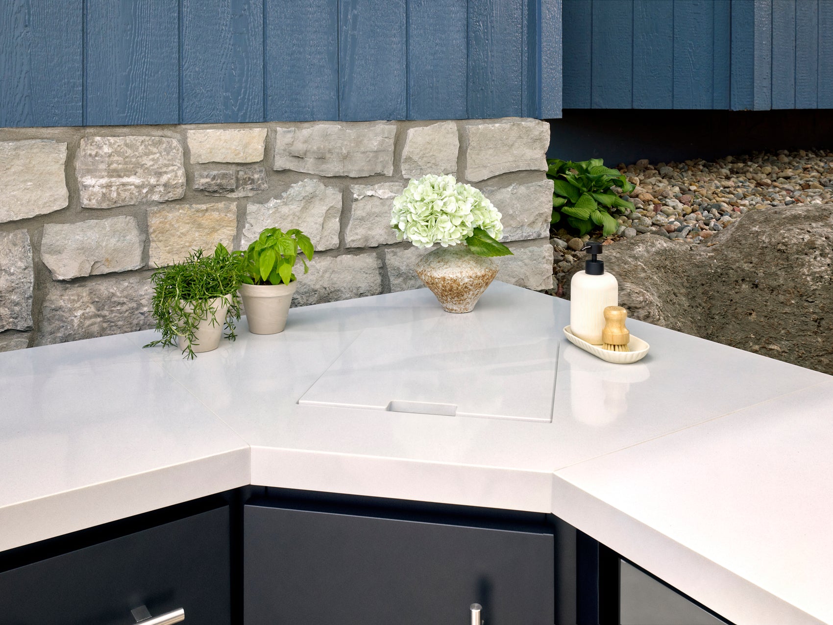Emory Outdoor kitchen sink built into white countertop with herbs, hydrangea, soap dispenser and brush nearby.