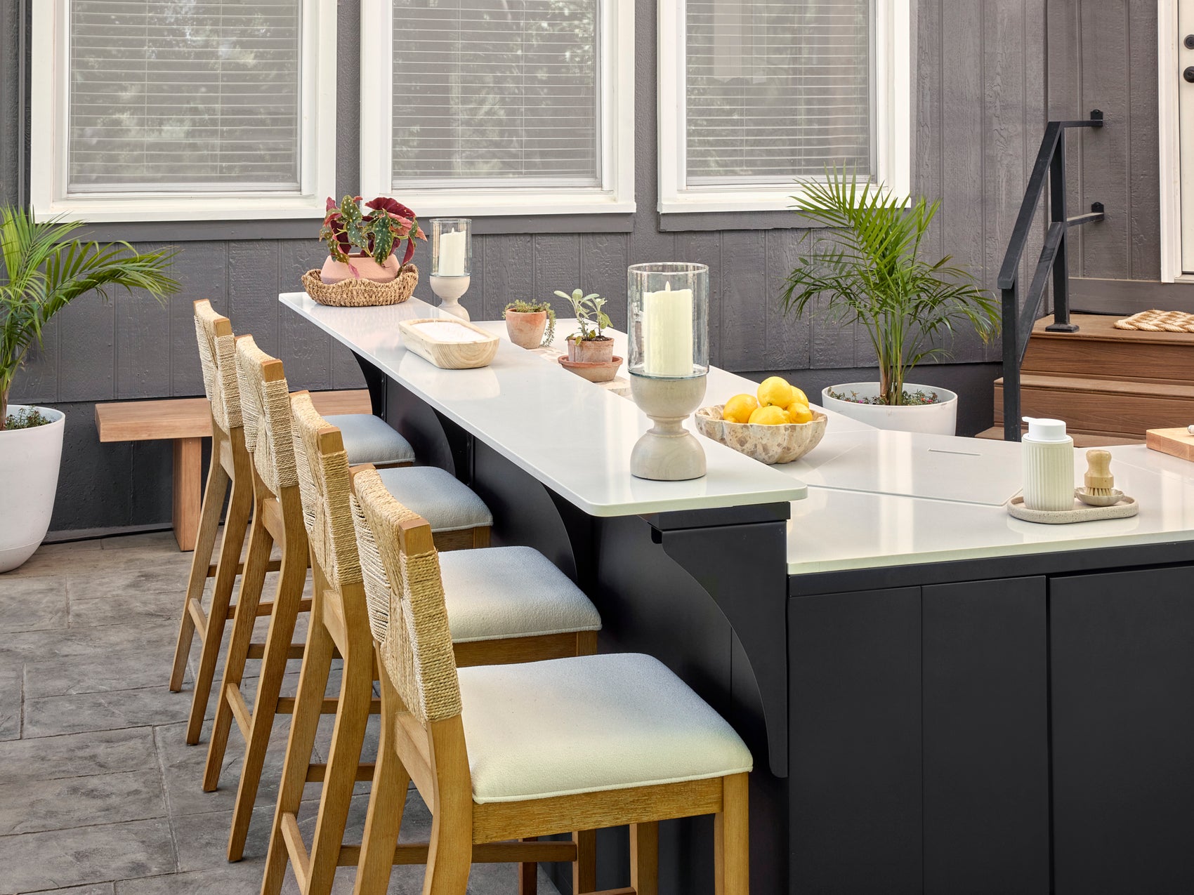 Outdoor patio bar with white engineered stone countertop, black cabinetry, and wooden bar stools with woven rope backs.