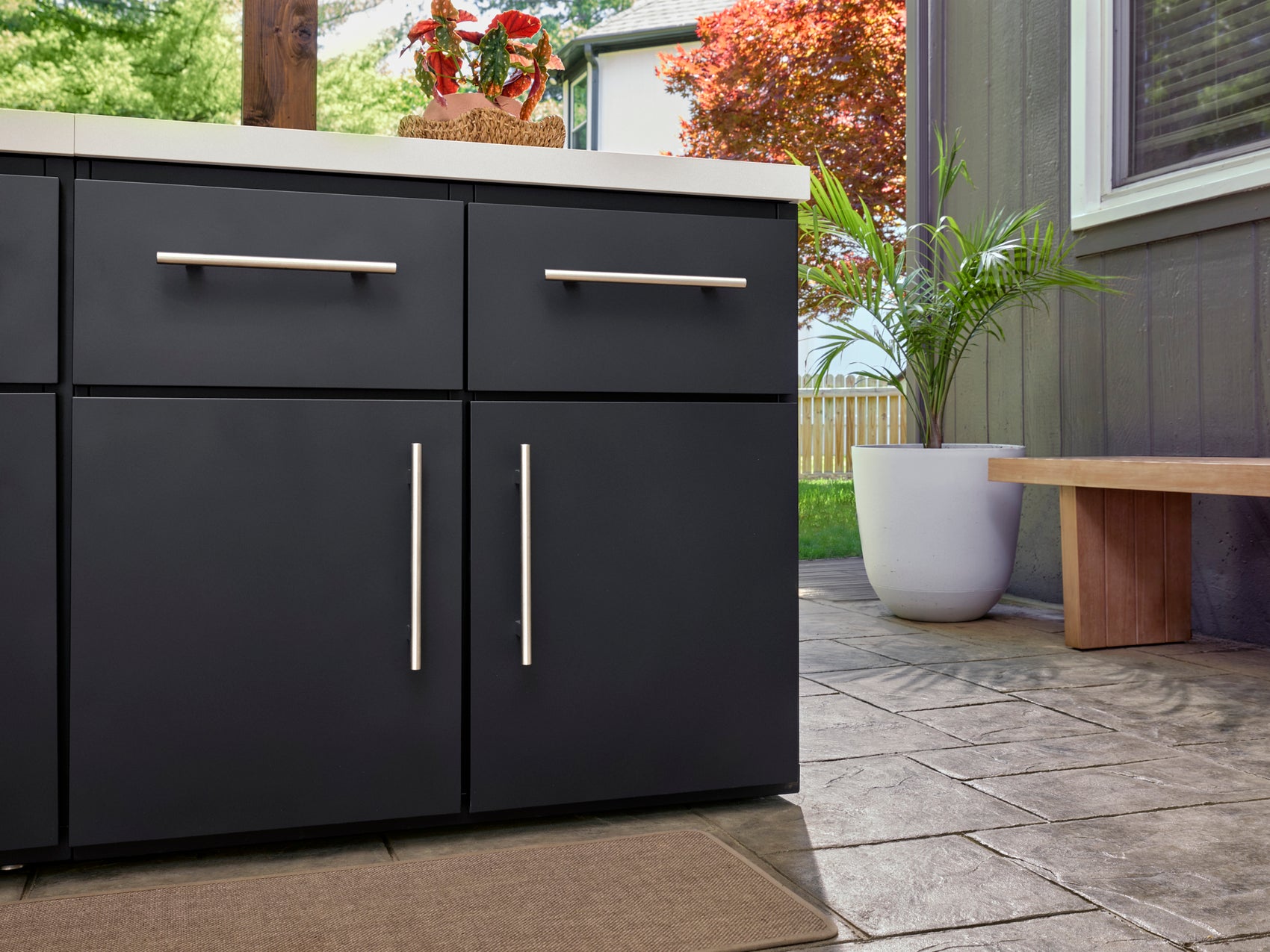 Matte black outdoor kitchen storage cabinets with metal bar pulls and white countertop on patio with potted palm.