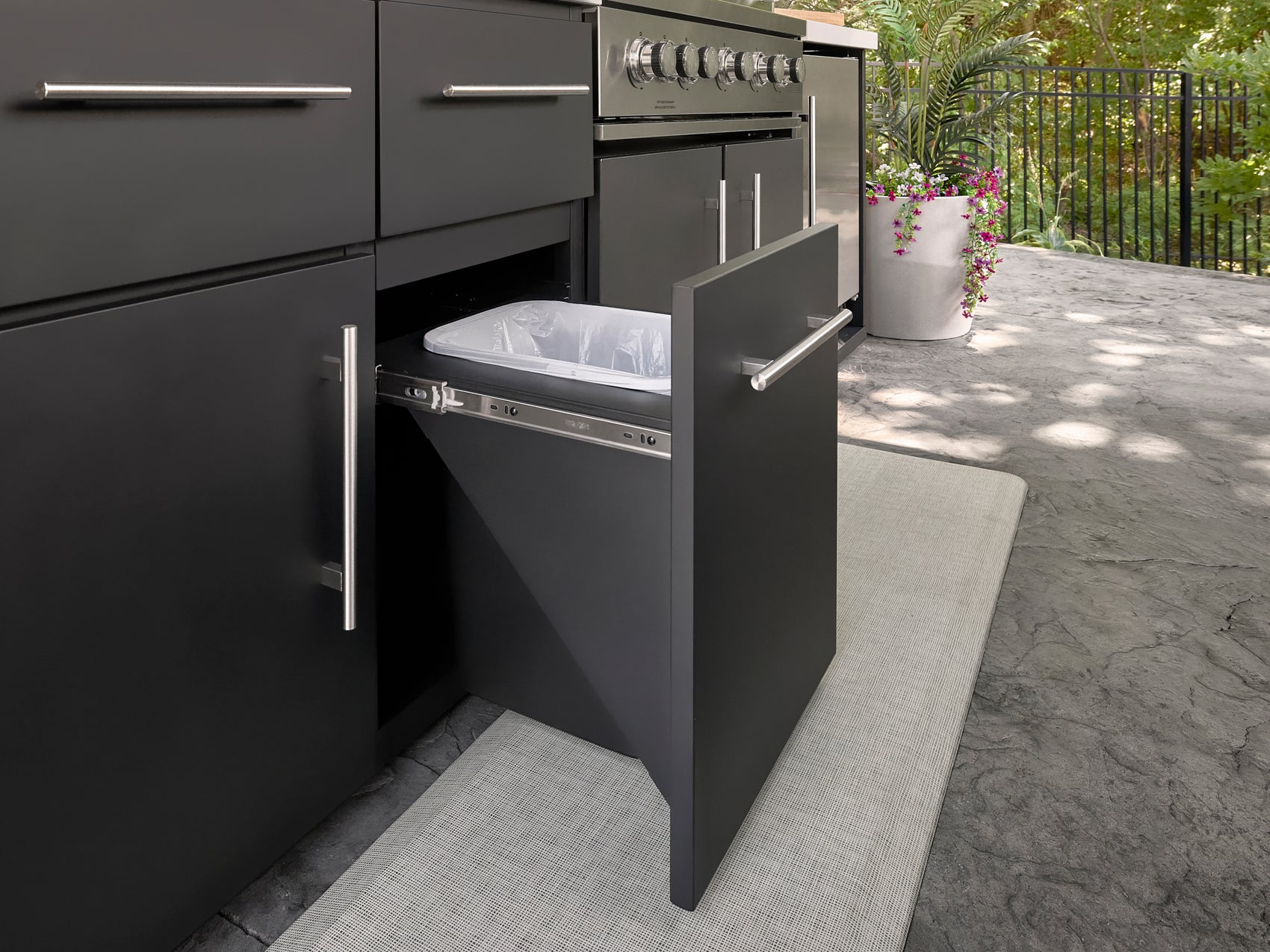 Pull-out trash bin beside stainless steel grill on covered kitchen with powder coated steel cabinetry.