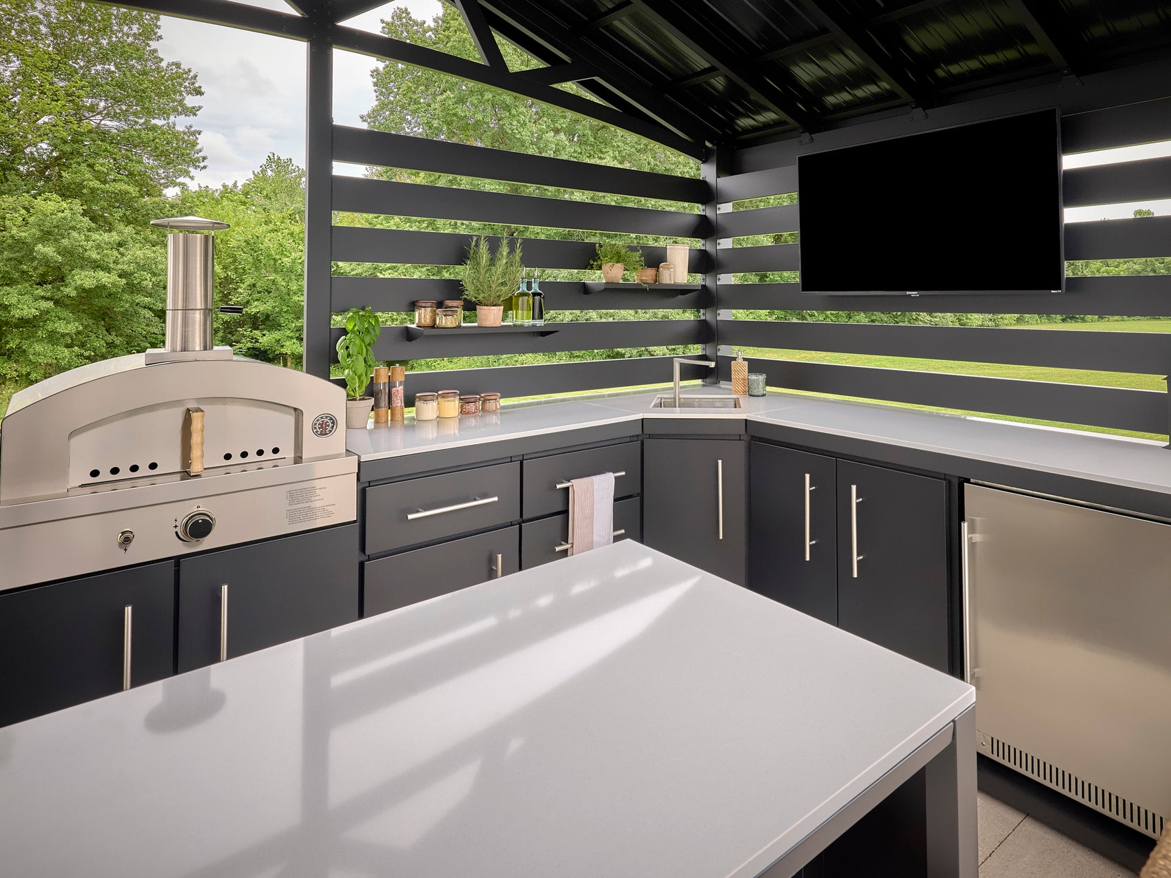 White outdoor countertop corner with dark gray cabinets, built-in pizza oven, sink, and horizontal slat privacy wall.