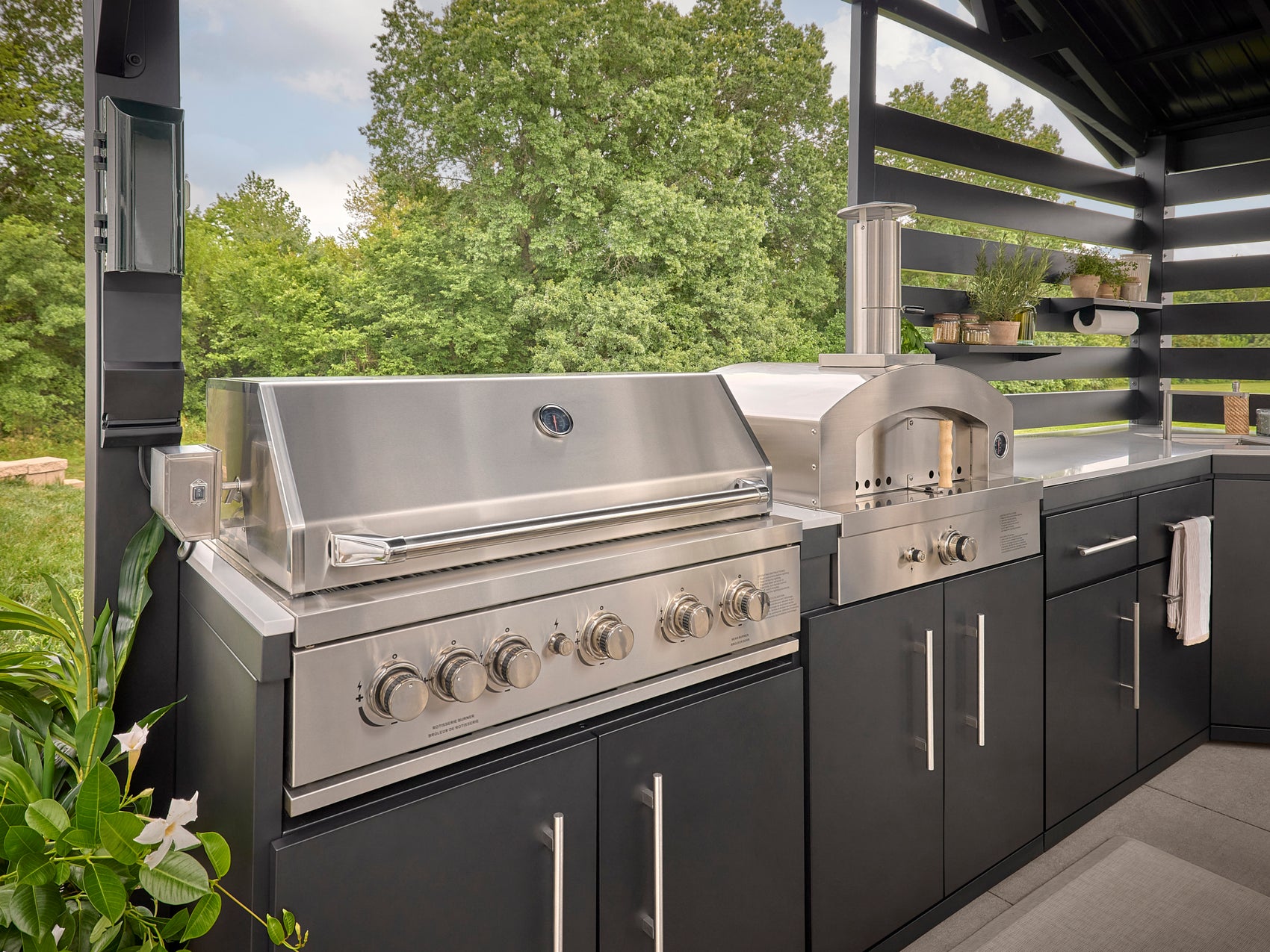 Built-in grill and pizza oven in outdoor cooking station with dark cabinetry and engineered stone countertops.