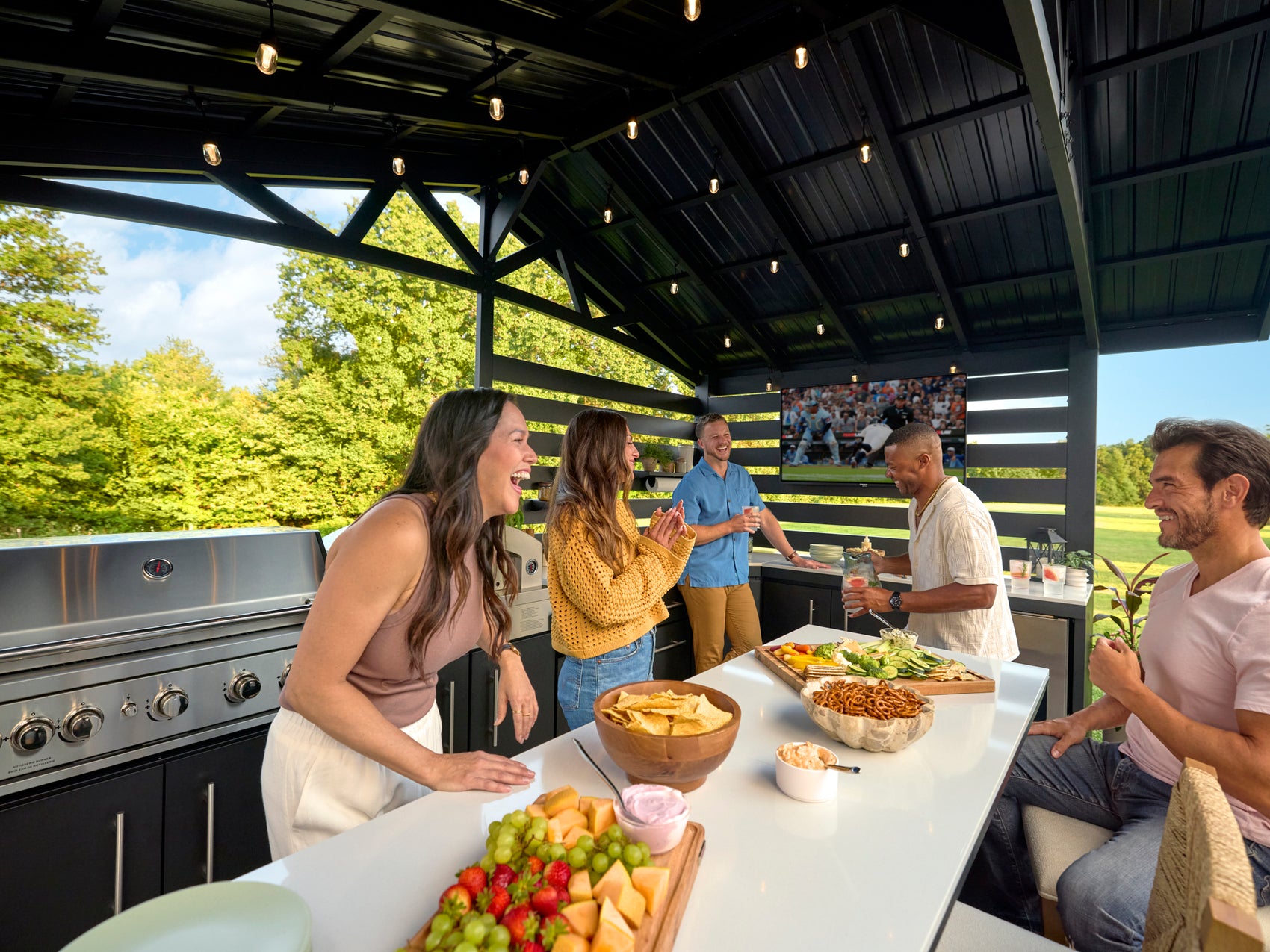 Friends gathered around the outdoor kitchen island under a black steel trellis roof with hanging Edison bulbs.