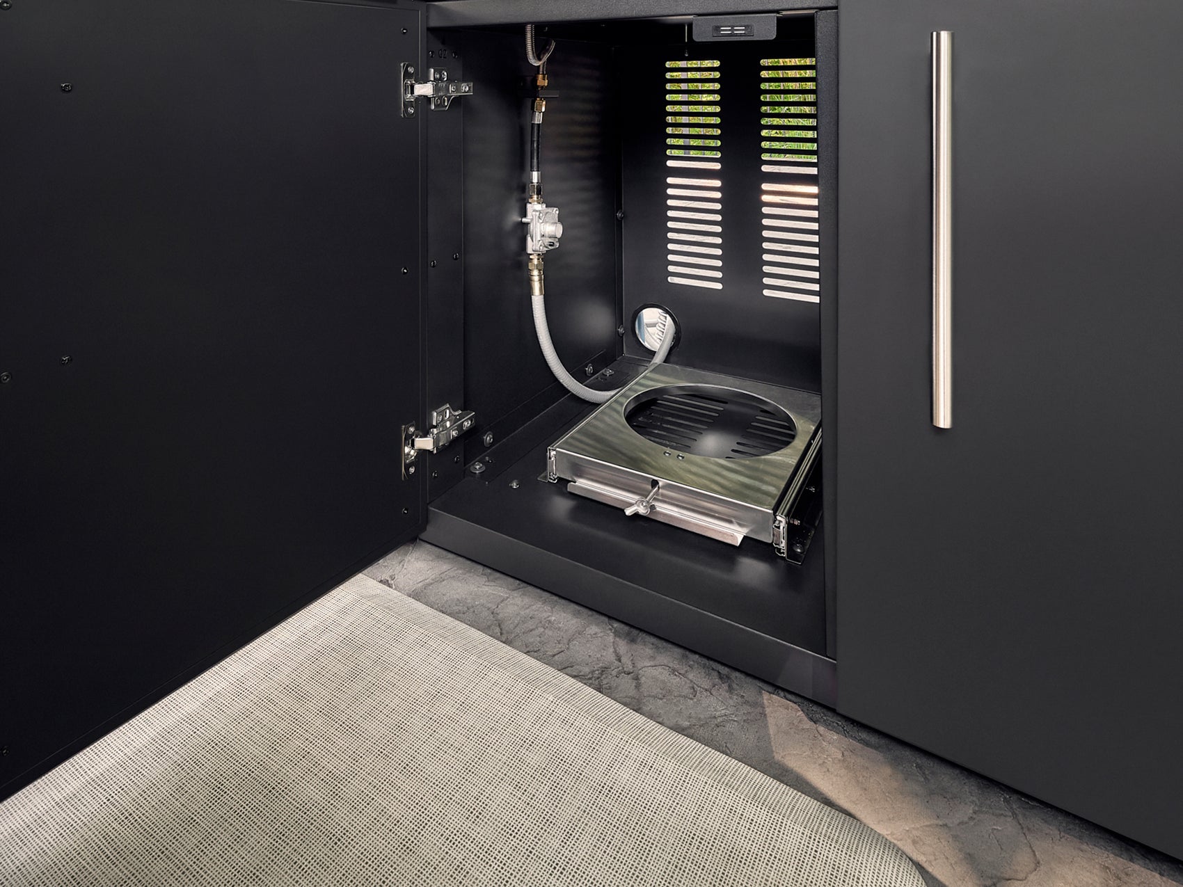 Black cabinet interior with built-in gas grill module, ventilation slots, and white supply line on gray tile floor.
