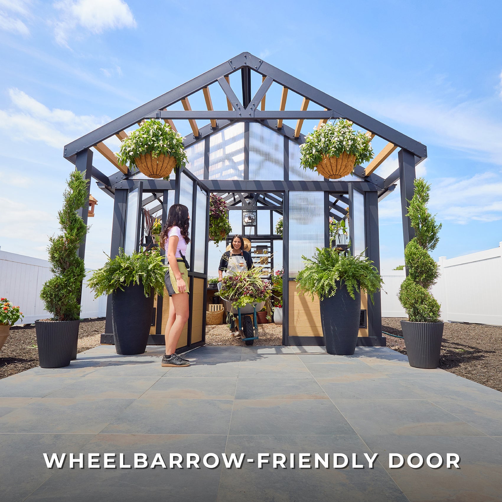 Two women use the wide entry door of the Larkspur walk-in greenhouse to roll a loaded wheelbarrow through it.