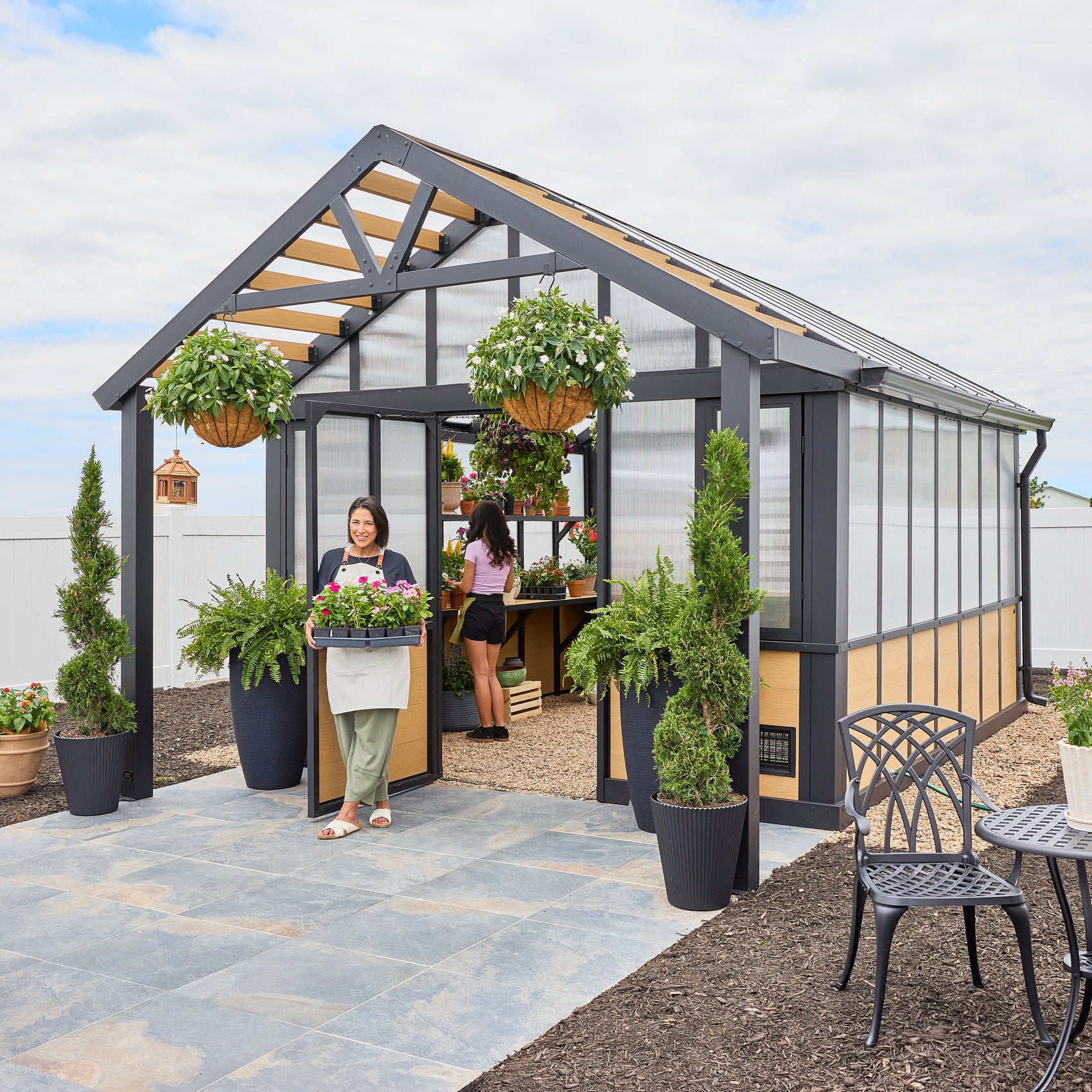 Backyard Discovery Larkspur outdoor greenhouse with two women tending potted plants and hanging flower baskets.