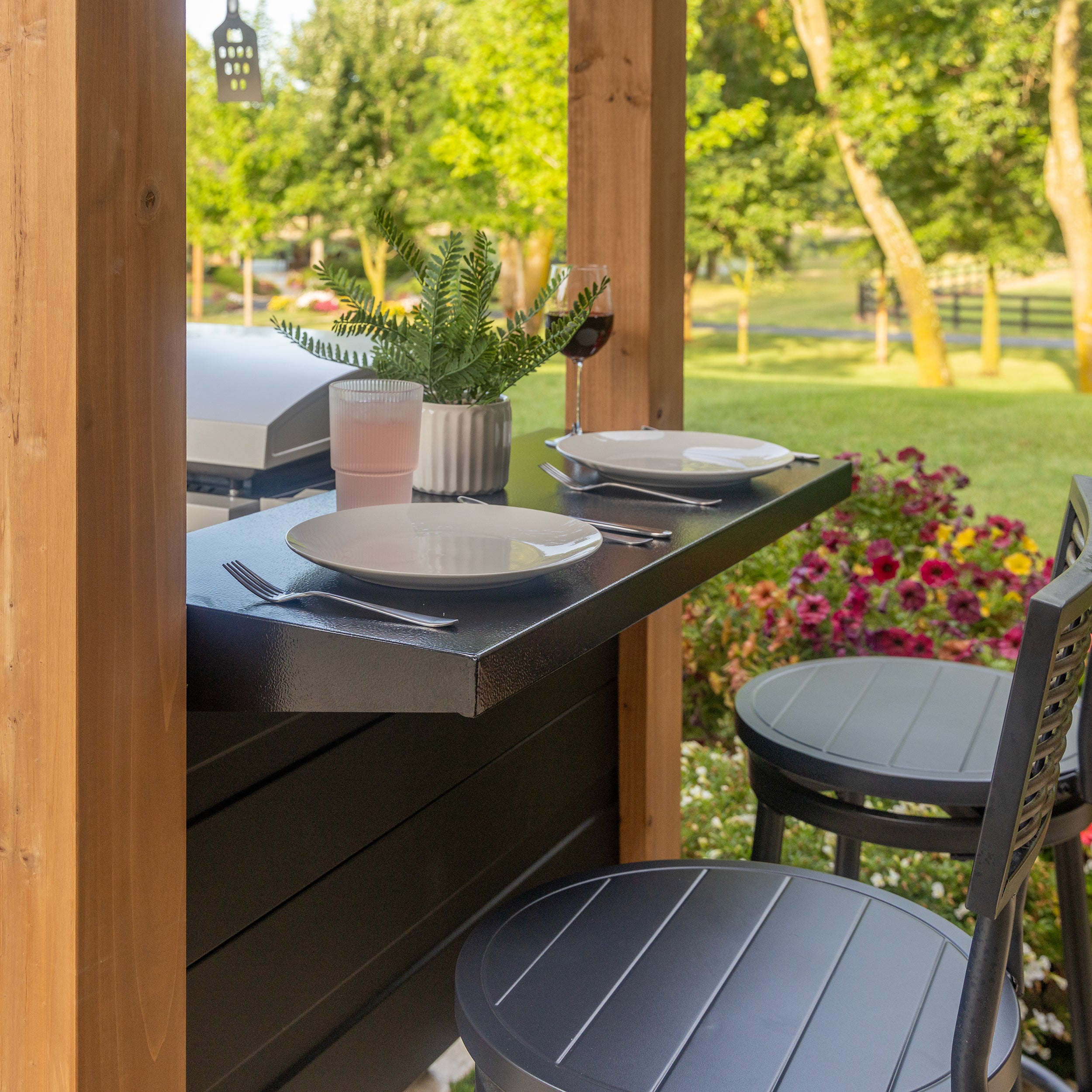 Wood grill gazebo with black steel countertops and place settings overlooking backyard garden.
