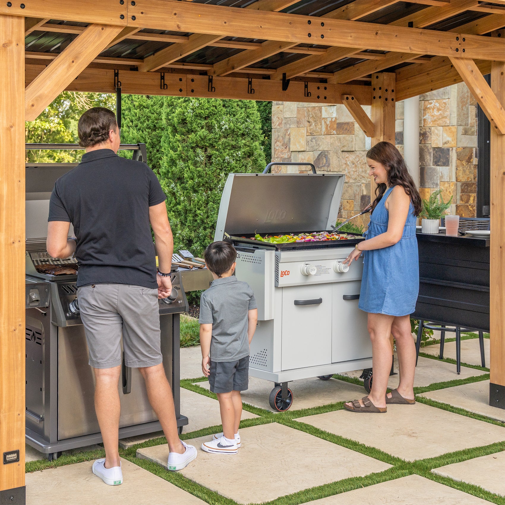Family grilling vegetables under cedar grill gazebo canopy with steel roof and countertops on patio.