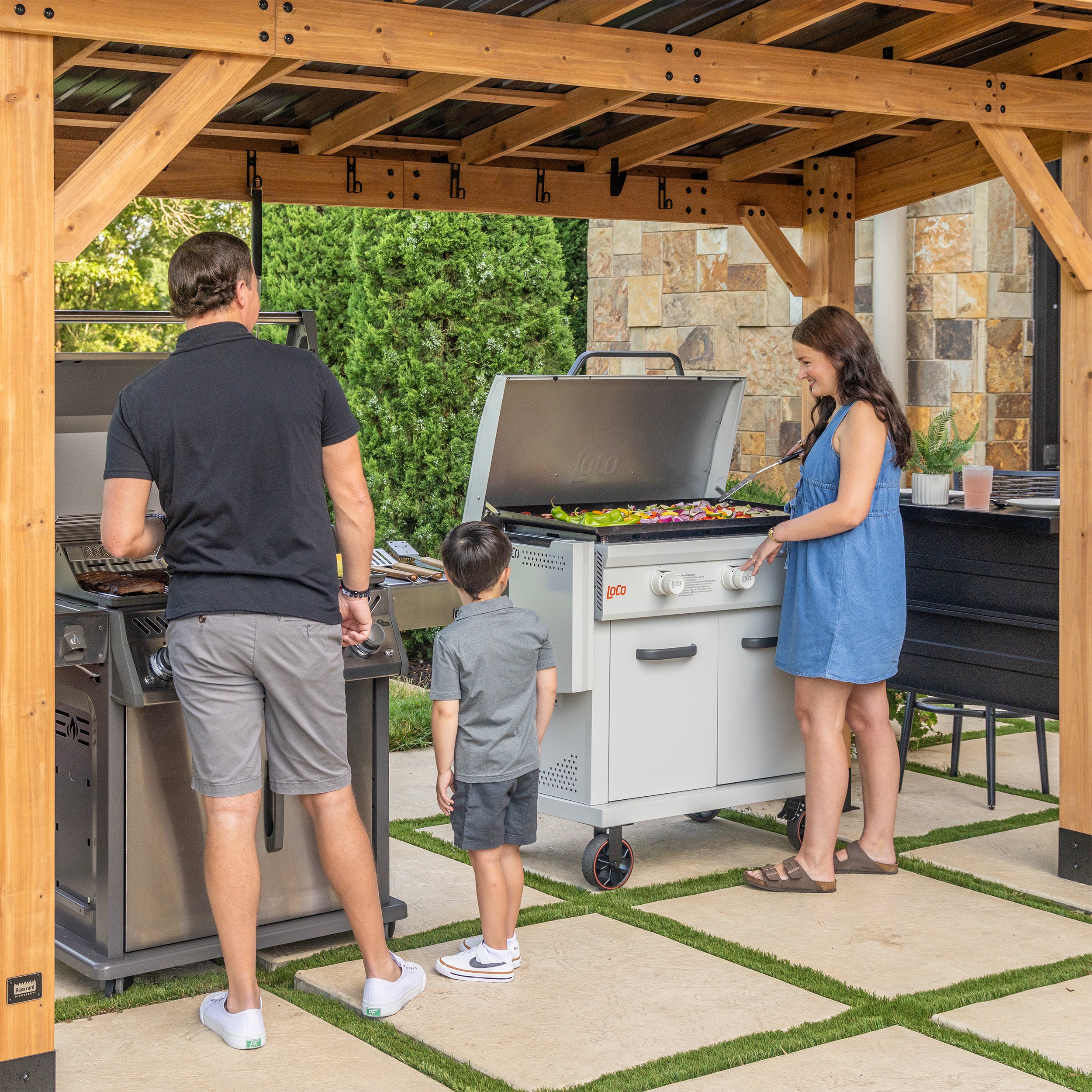 Family grilling vegetables under cedar grill gazebo canopy with steel roof and countertops on patio.
