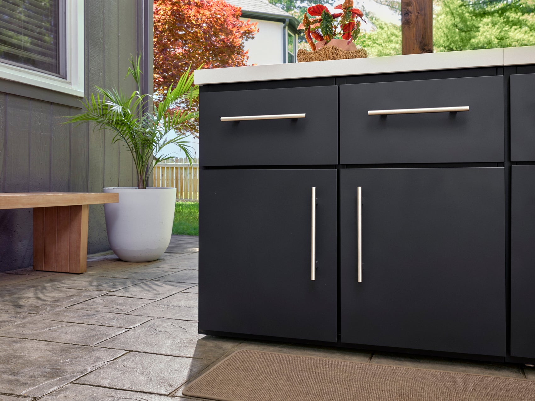 Black powder-coated Emory outdoor cabinet with brushed metal handles and drawers on a patio with stone pavers and greenery.
