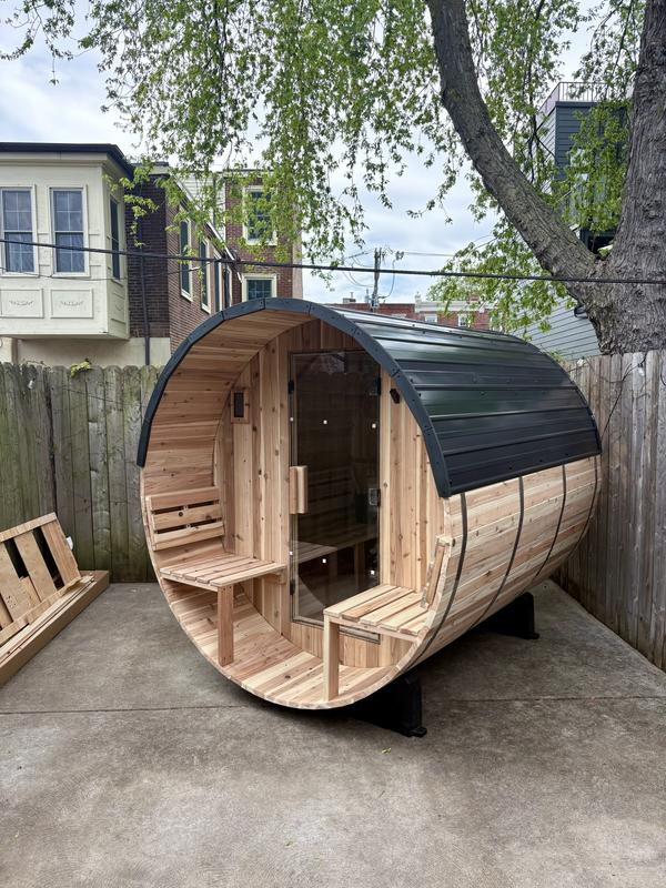 Wooden barrel-shaped sauna in a backyard setting with trees and houses in the background.