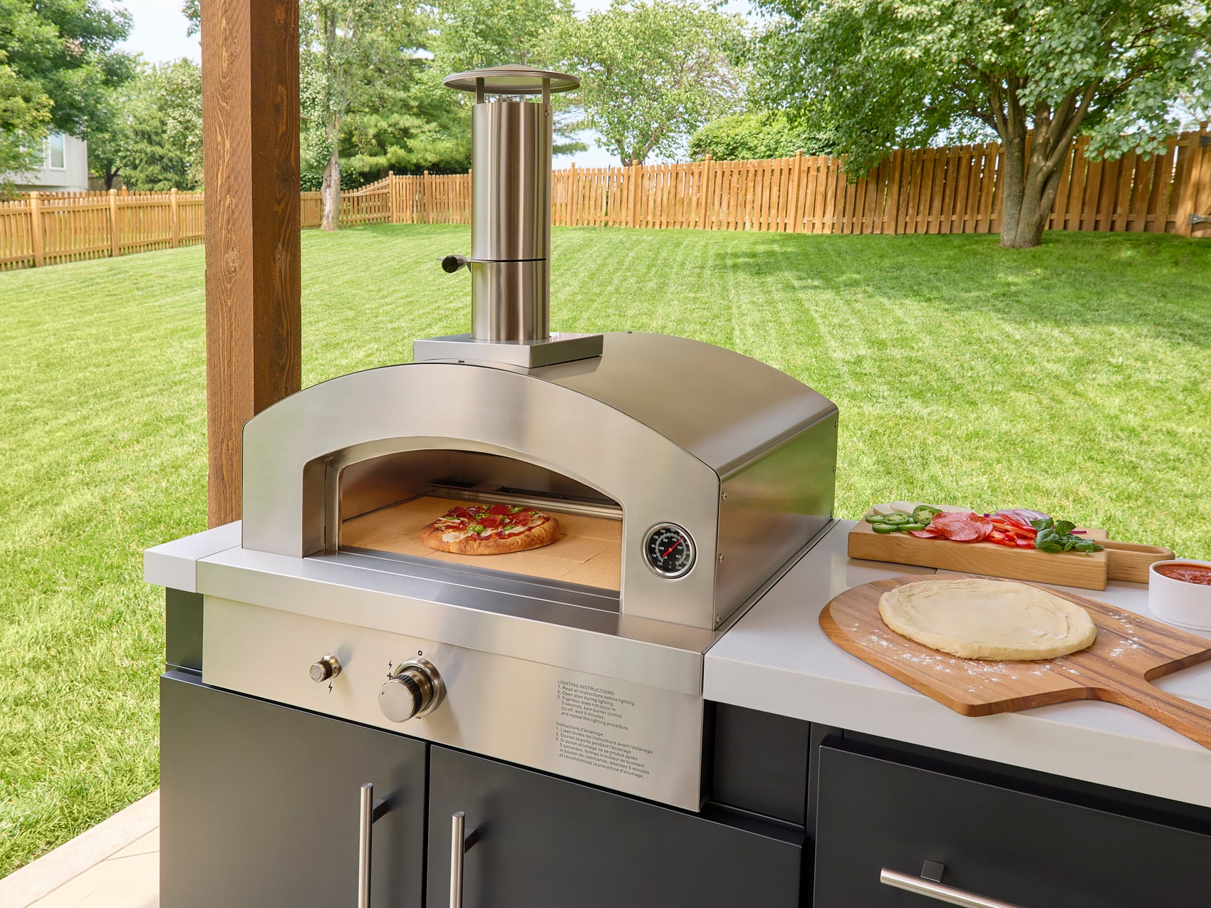 Outdoor pizza oven with arched opening, temperature gauge, and chimney on countertop with pizza and prep ingredients.