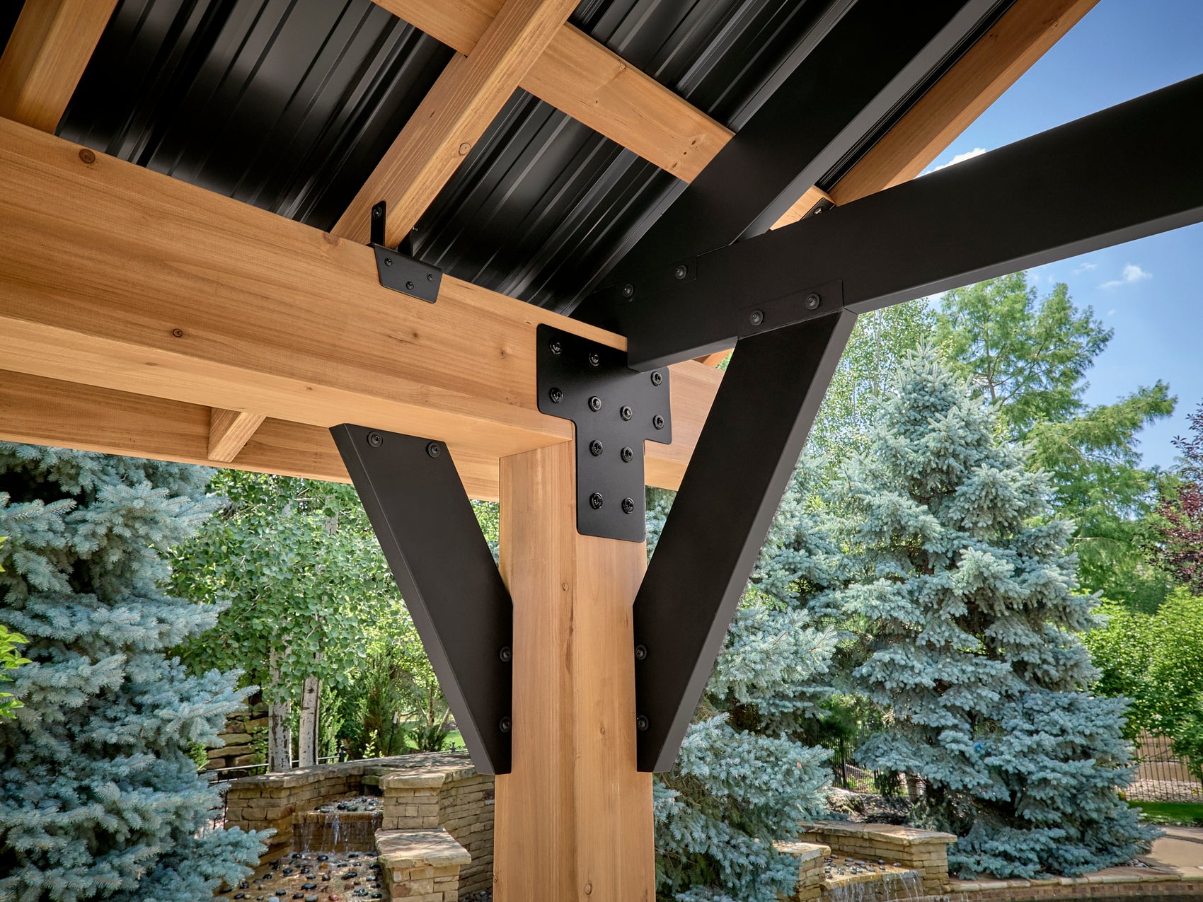 Black steel corner bracket connecting wooden post and beam on Lodge outdoor gazebo with metal roof and garden view.