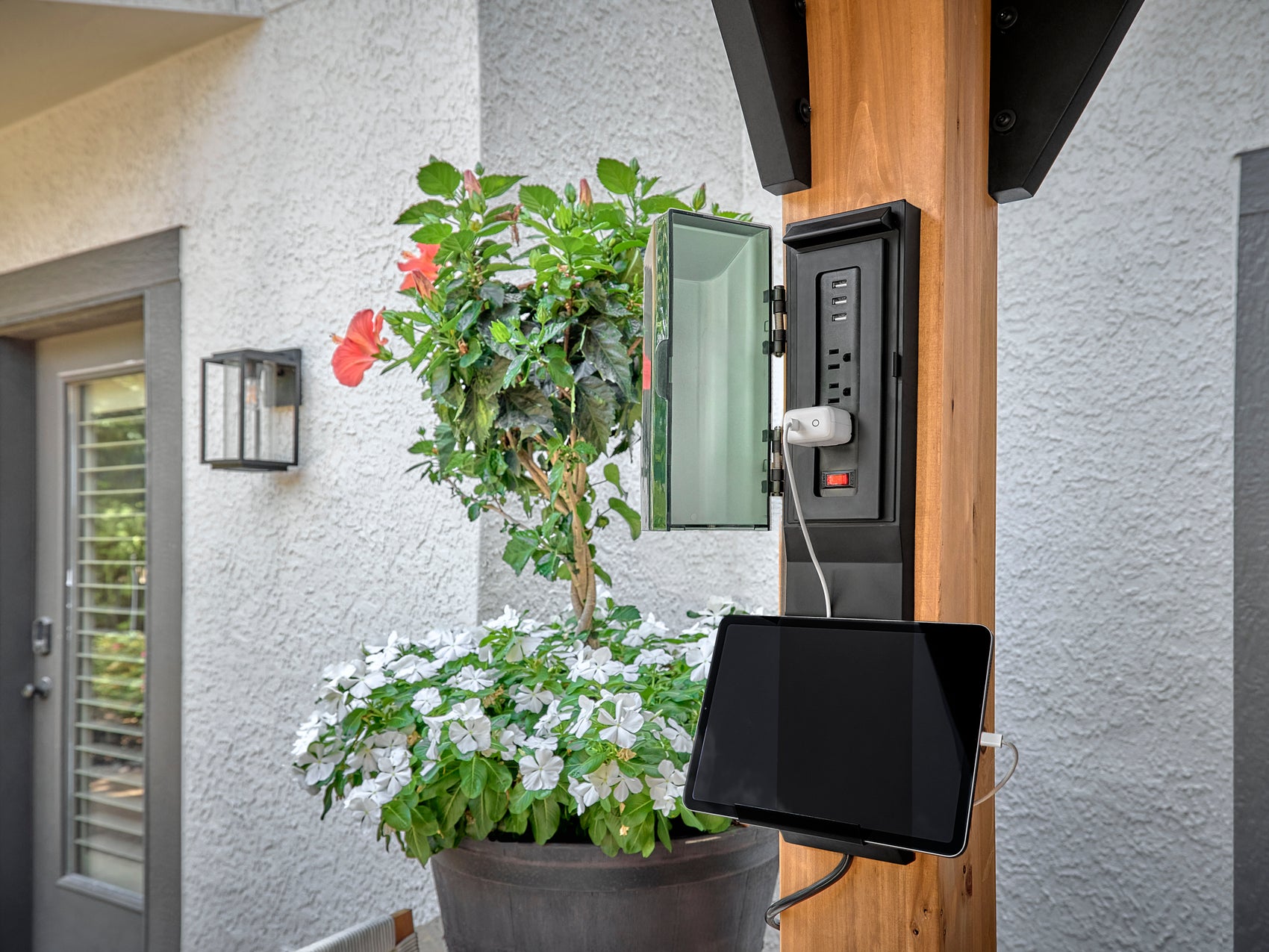 Outdoor power outlet box with USB ports and mounted tablet on steel Lodge gazebo hardware post beside flowering plants.